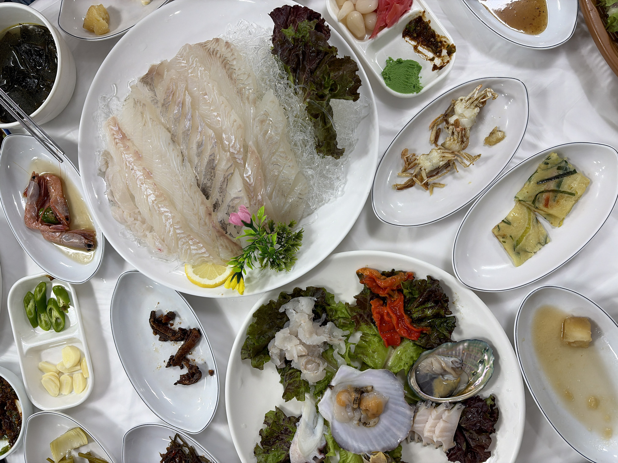 The image shows a spread of Korean food on a white tablecloth, likely in a restaurant in Toseong-myeon, South Korea. The dishes include a large platter of thinly sliced raw fish, a plate of assorted seafood such as scallops and abalone, a small bowl of soup, a plate with a cooked shrimp, small side dishes of pickled garlic and peppers, dried seasoned vegetables, fried seafood pancakes, and a dish with a cooked crab. There are also small dishes containing sauces and condiments like wasabi and pickled ginger. Chopsticks are visible in the soup bowl. The setting appears to be a mealtime gathering.