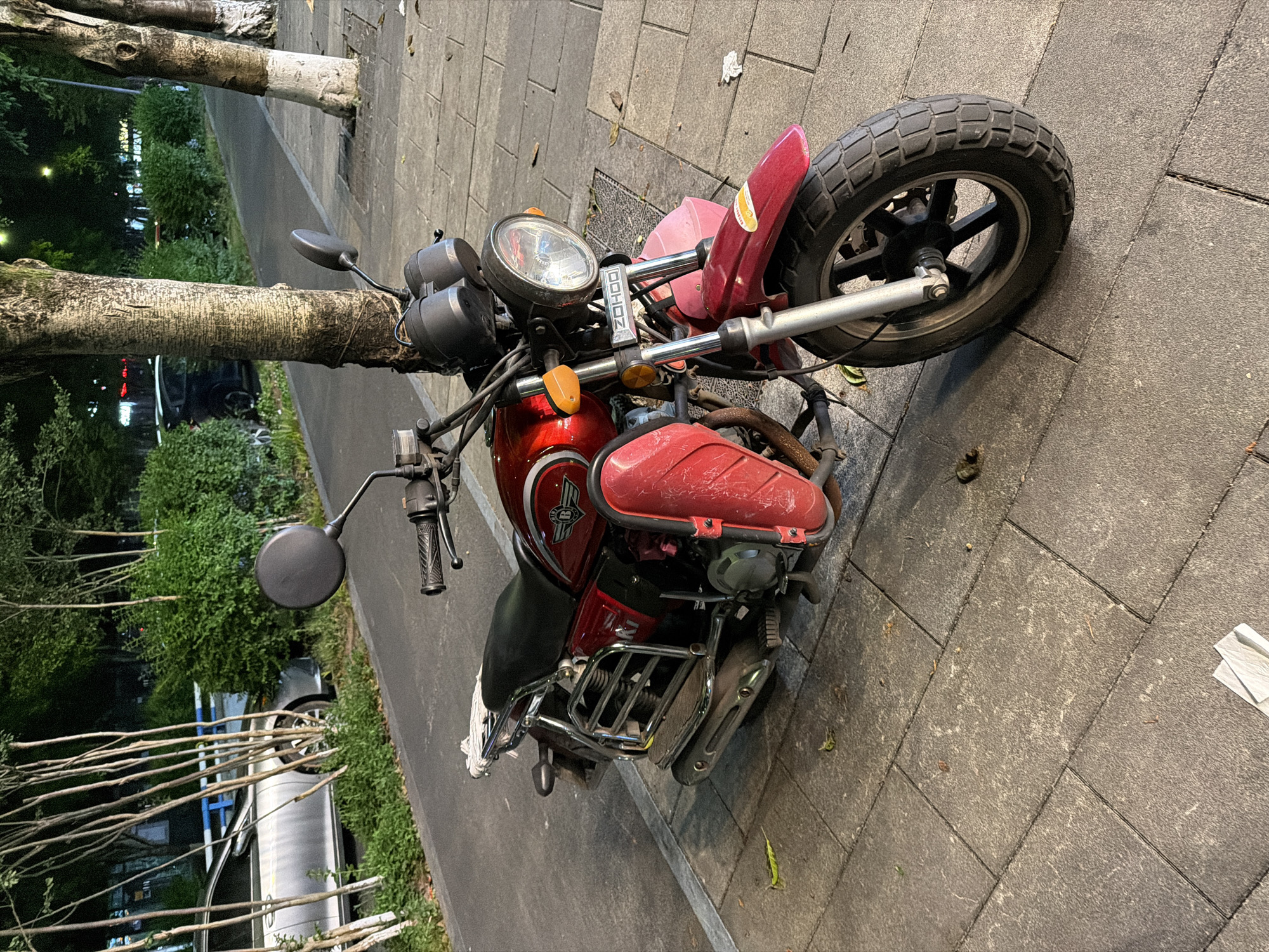 A red and black Zhejiang ZongShen motorcycle is parked on a paved sidewalk in Chongqing High-tech Zone, China. The motorcycle is situated adjacent to a row of trees lining the walkway. The lighting suggests it is either late evening or early morning. The environment appears urban and landscaped.