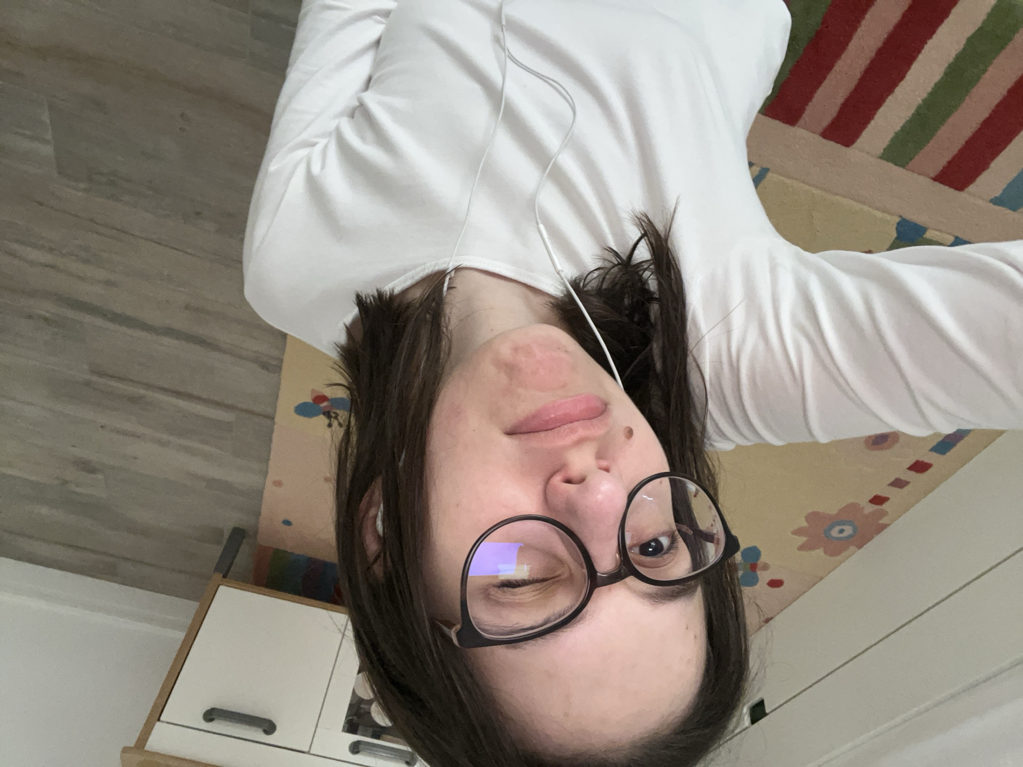 A casually oriented indoor selfie depicts a young individual, likely female, lying on their back, looking towards the camera. The person, positioned upside down in the frame, has dark hair and is wearing a white long-sleeved shirt. Black-framed glasses are worn, and white wired earphones are draped around their neck and chest. One eye is open, while the other is closed in a distinct wink, conveying a relaxed and playful expression. There is visible redness or skin texture around the chin and cheek area.
The background shows elements of a residential interior. To the left, light-toned wooden laminate flooring is visible. Below the person, a beige rug with subtle, possibly floral or abstract patterns can be seen. Another colorful rug, featuring prominent red, green, and blue stripes on a lighter base, is partially visible towards the top right, behind the person's arm. In the bottom left corner, part of a light-colored cabinet or shelf with white doors and dark handles is present. The scene captures a private moment of relaxation within what is contextually specified as Marten, Bulgaria. No visible text is present in the image.