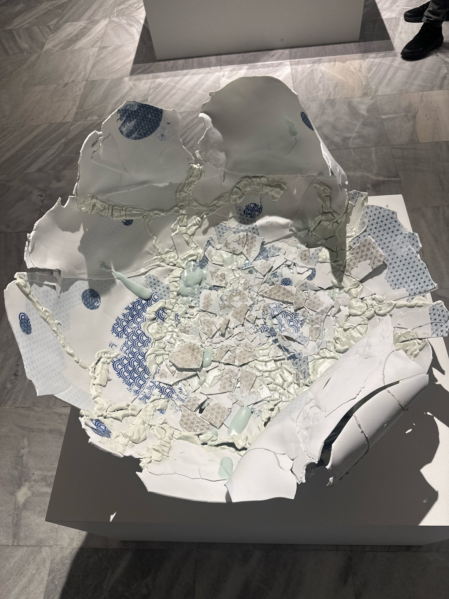 The image shows a large, broken ceramic sculpture displayed on a white pedestal in what appears to be an art gallery or exhibition space. The sculpture is predominantly white, with fragments of blue and beige patterns, possibly traditional Japanese motifs like waves and geometric shapes. The ceramic pieces are held together by white and light green adhesive or molding material, creating a textured and fragmented appearance. The sculpture is centrally located in the frame, with a marble-tiled floor in the background, exhibiting a subtle grey veining. Lighting from an unseen source casts shadows across the sculpture and the floor. In the upper right corner of the image, the lower legs and black shoes of at least one person are visible, suggesting they are standing and observing the artwork. The overall setting implies a contemporary art exhibition. The location is specified as Belgrade, Serbia.