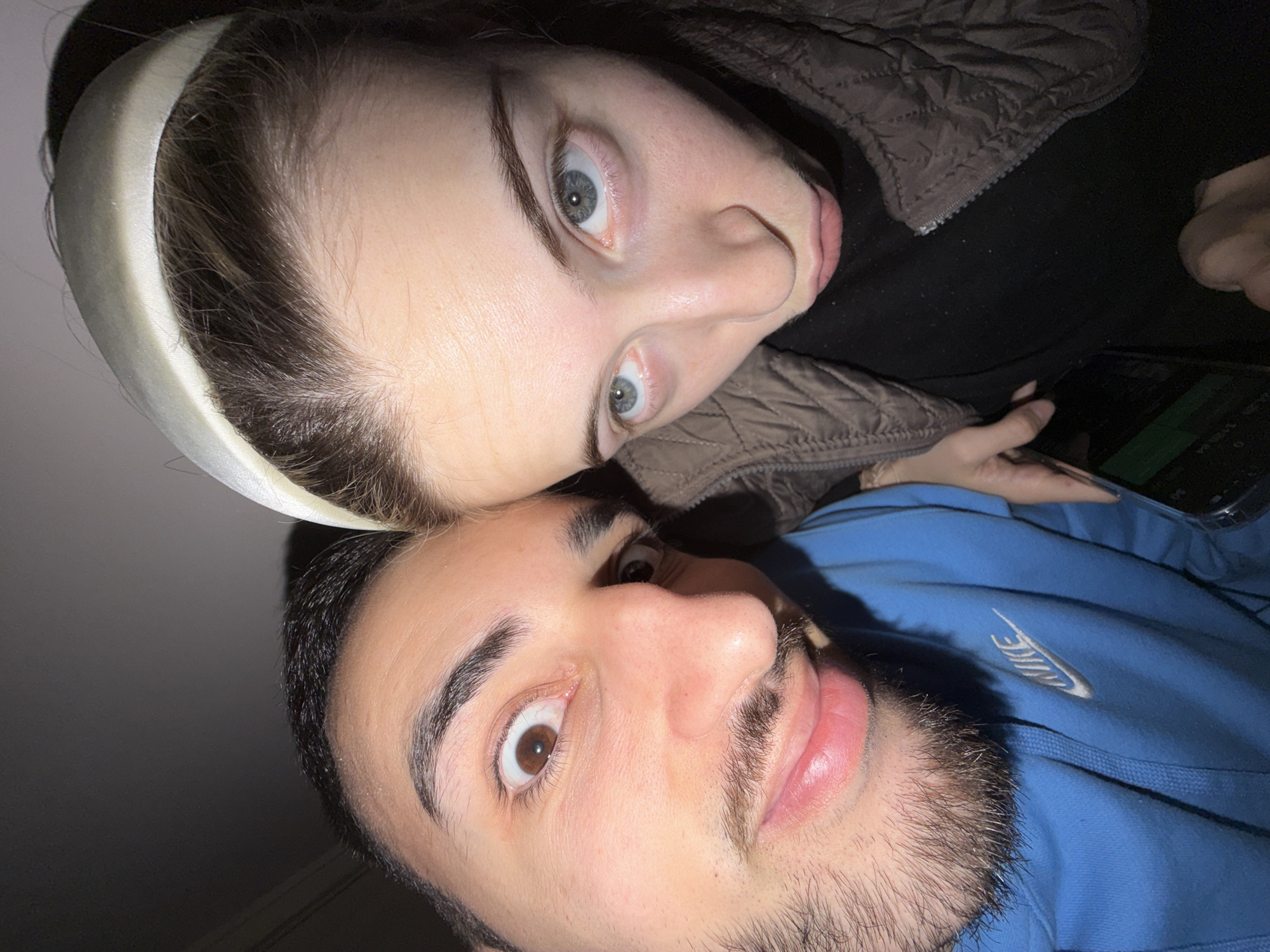 A close-up selfie captures two individuals. The person on the bottom is male, with brown eyes, dark hair, and a short beard. He wears a blue Nike hoodie. Above him is a female with light grey eyes and a white headband. She wears a quilted jacket with a black top. In the background, a hand is visible holding a phone. The location in the city of Milevo, Bulgaria cannot be determined from this image.