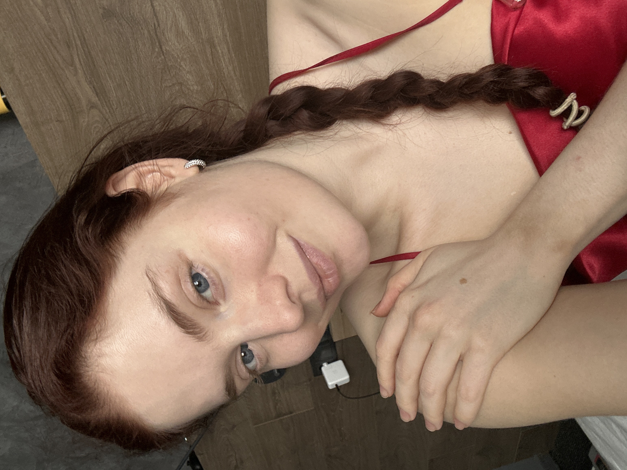 A woman with light skin, auburn hair styled in a braid, and blue eyes is positioned at an angle in the frame, against what appears to be a wooden surface. She is wearing a red top. The image includes an ear cuff on her left ear and a hair clip at the end of her braid. A charging block with a cord is visible in the background. The environment is unspecified, and there is no indication of the location being Riga, Latvia.