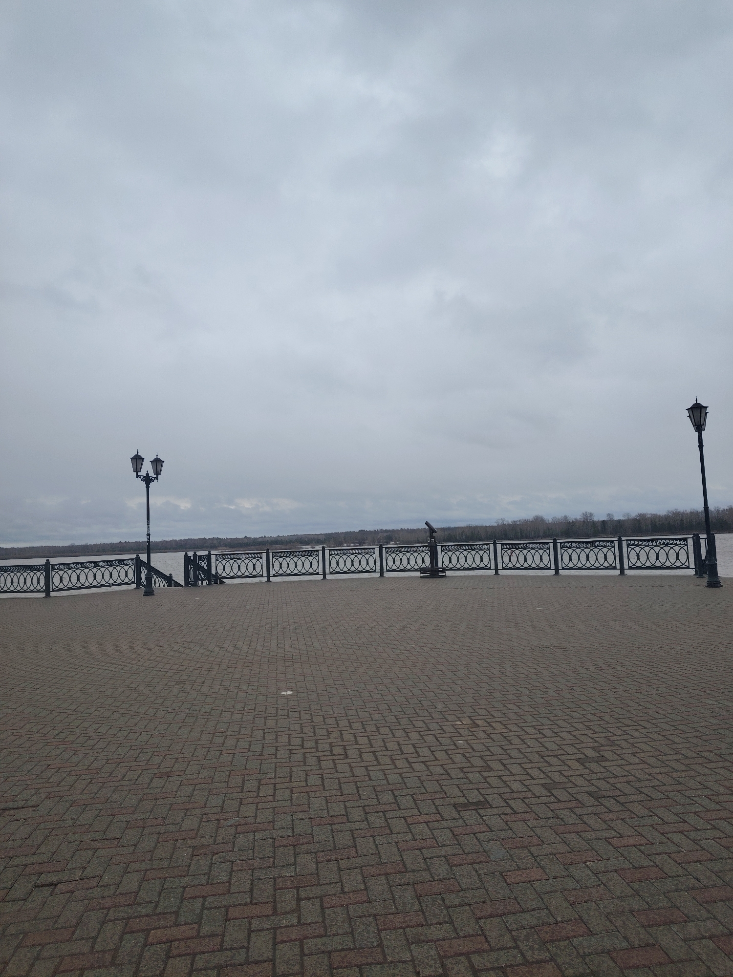 The image depicts a wide, brick-paved promenade in Sarapul, Russia, likely along the Kama River. The sky is overcast. A black wrought-iron fence runs along the edge of the promenade, separating it from the river. Two decorative lampposts stand along the promenade, one with a double light fixture and another with a single. A viewing telescope is mounted on a stand along the fence. The river and a distant treeline are visible beyond the fence. The promenade is empty of people.