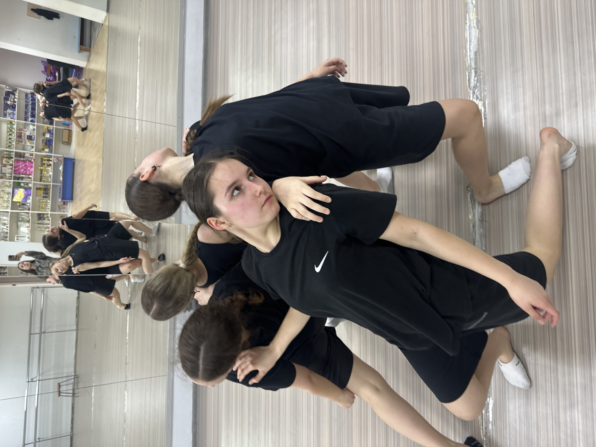 A group of young women are in a dance studio in Votkinsk, Russia. They are wearing black t-shirts and shorts. The woman in the foreground is wearing a black Nike t-shirt and looking up and to the left. Another woman is behind her, with her hand on the first woman's shoulder. The background of the image shows a mirror reflecting the group, as well as shelves stocked with items. The floor is a light-colored wood.