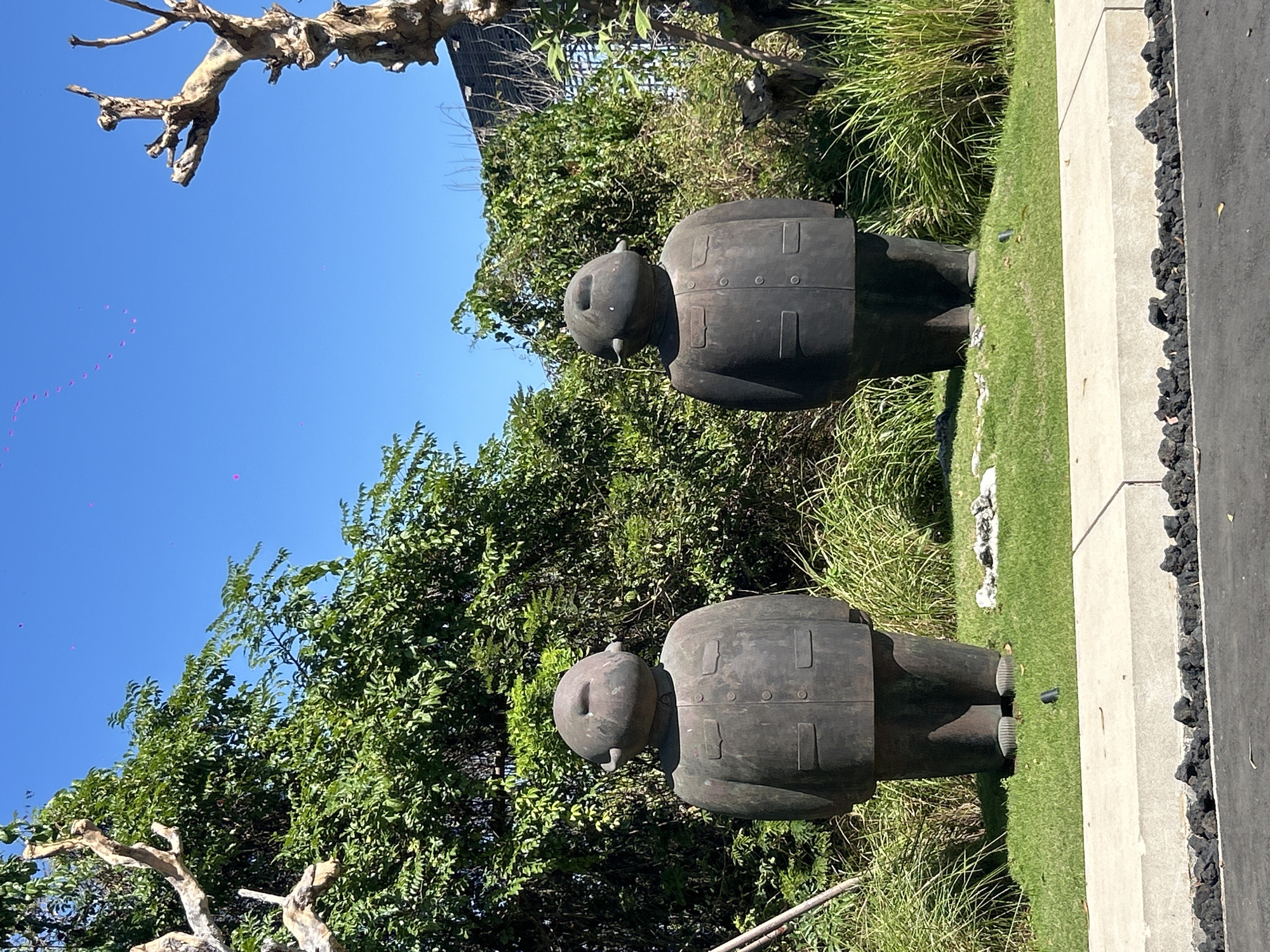 The image depicts two large, dark-colored, abstract statues resembling figures in suits, standing on a manicured lawn. The statues are positioned outdoors in what appears to be a garden or park setting. They are dark, roughly human-shaped, with large heads, small ears, and rounded bodies. The figures are dressed in what resembles jackets with buttons and pockets.
Behind the statues, lush green foliage and trees form a dense backdrop. The sky above is a clear, vibrant blue. To the left and right of the frame, the bare, gnarled branches of dead trees are visible, contrasting with the living greenery. The foreground shows a paved area with a concrete border, and beyond that, a darker surface, possibly asphalt.
The statues are placed on a neatly mowed lawn, with some scattered white rocks or debris around their bases. The overall impression is one of an outdoor art installation. The scene is well-lit by sunlight, casting shadows on the statues and the ground. The atmosphere is calm and serene, with the bright blue sky and green vegetation creating a pleasant, natural environment. The location is stated to be in the city of Kuta Selatan, Indonesia.
