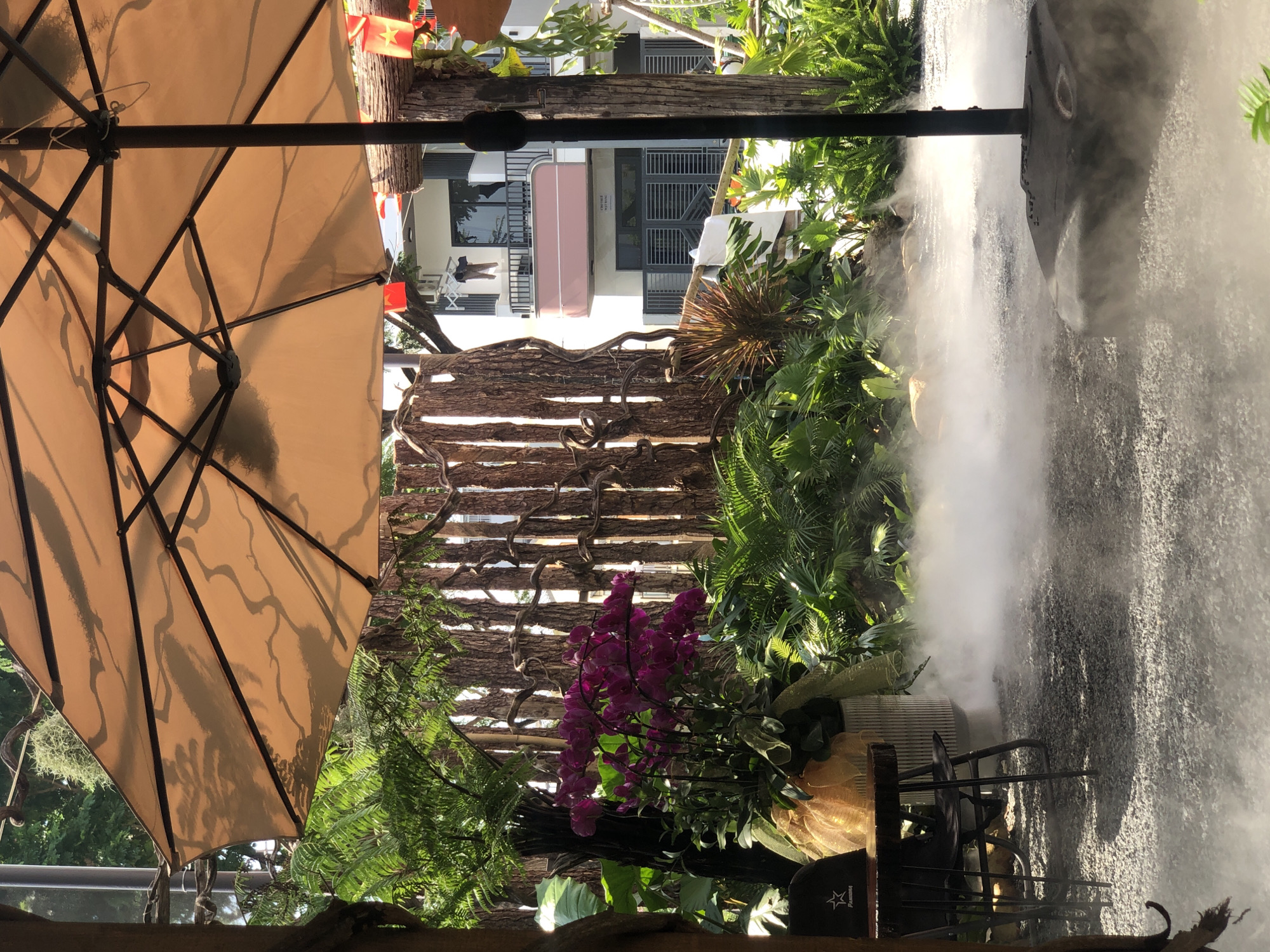 The image shows an outdoor café scene in Đà Nẵng, Vietnam. A large beige umbrella occupies the upper left quadrant. A decorative wooden fence with intertwined branches is partially visible behind a lush arrangement of ferns and a vibrant bunch of purple orchids. A water feature or artificial waterfall creates a misty environment to the right. Through gaps in the wooden fence, glimpses of a modern building can be seen in the background. The image is taken from a low angle, looking upward.