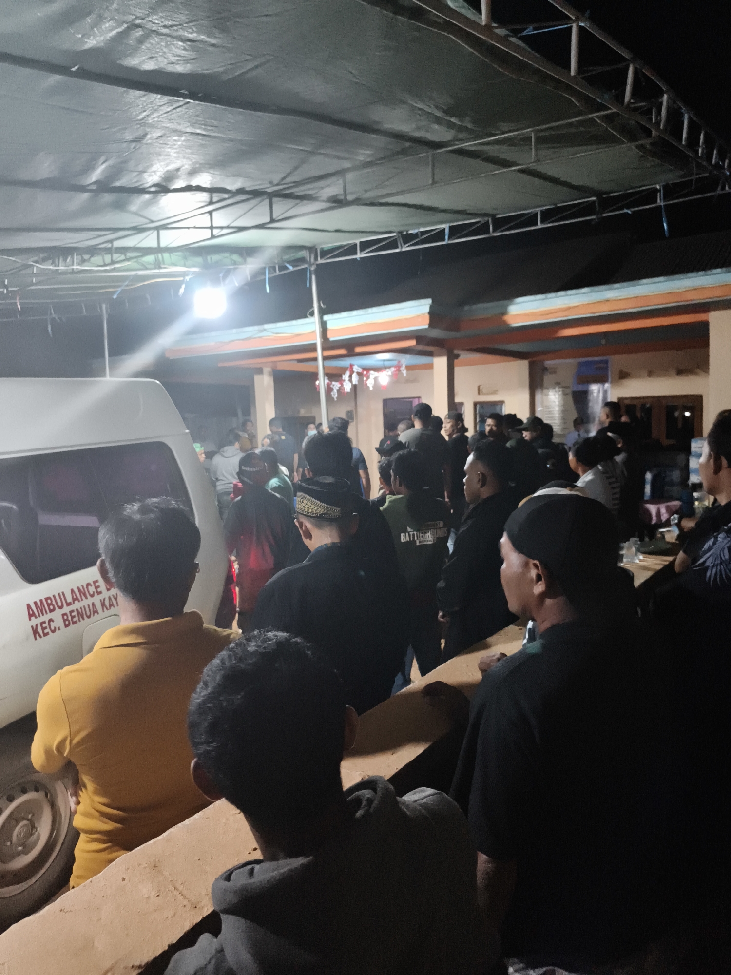 The image depicts a nighttime scene outside what appears to be a community building or hall in the city of Selimpatan Jaya, Indonesia. A white ambulance with the text "AMBULANCE KEC. BENUA KAY" is parked on the left side of the frame, its lights illuminating the area. A crowd of people, predominantly men, are gathered around the ambulance and the building. Many are wearing dark clothing, with some sporting traditional headwear. The atmosphere seems somber or expectant. The environment includes a large, dark tarp stretched overhead, supported by metal framing, suggesting an outdoor gathering or shelter. The building in the background is lit and appears to be a simple structure with visible windows and an orange and blue trim. The lighting is artificial and somewhat harsh, casting shadows.