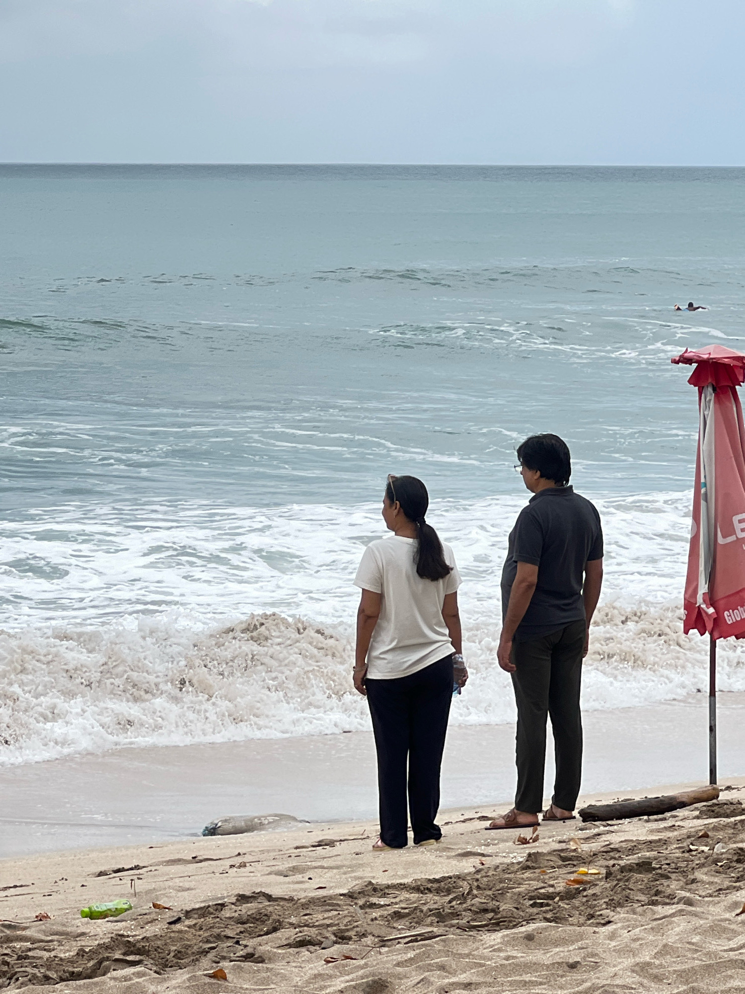 Two individuals stand on a sandy beach in Kuta, Indonesia, facing the ocean. The person on the left, viewed from behind, wears a white t-shirt and dark pants, with their hair pulled back in a ponytail. The person on the right, also viewed from behind, is wearing a dark t-shirt and dark pants. Both are looking out at the water. Waves are breaking on the shore. In the distance, a lone surfer is visible in the water. To the right of the frame, a red umbrella is partially visible. The sky is overcast. The sand is a light brown color, with some debris, including a green plastic bottle and leaves, scattered on the surface. A piece of driftwood lies on the sand. The sea appears calm with gentle waves.