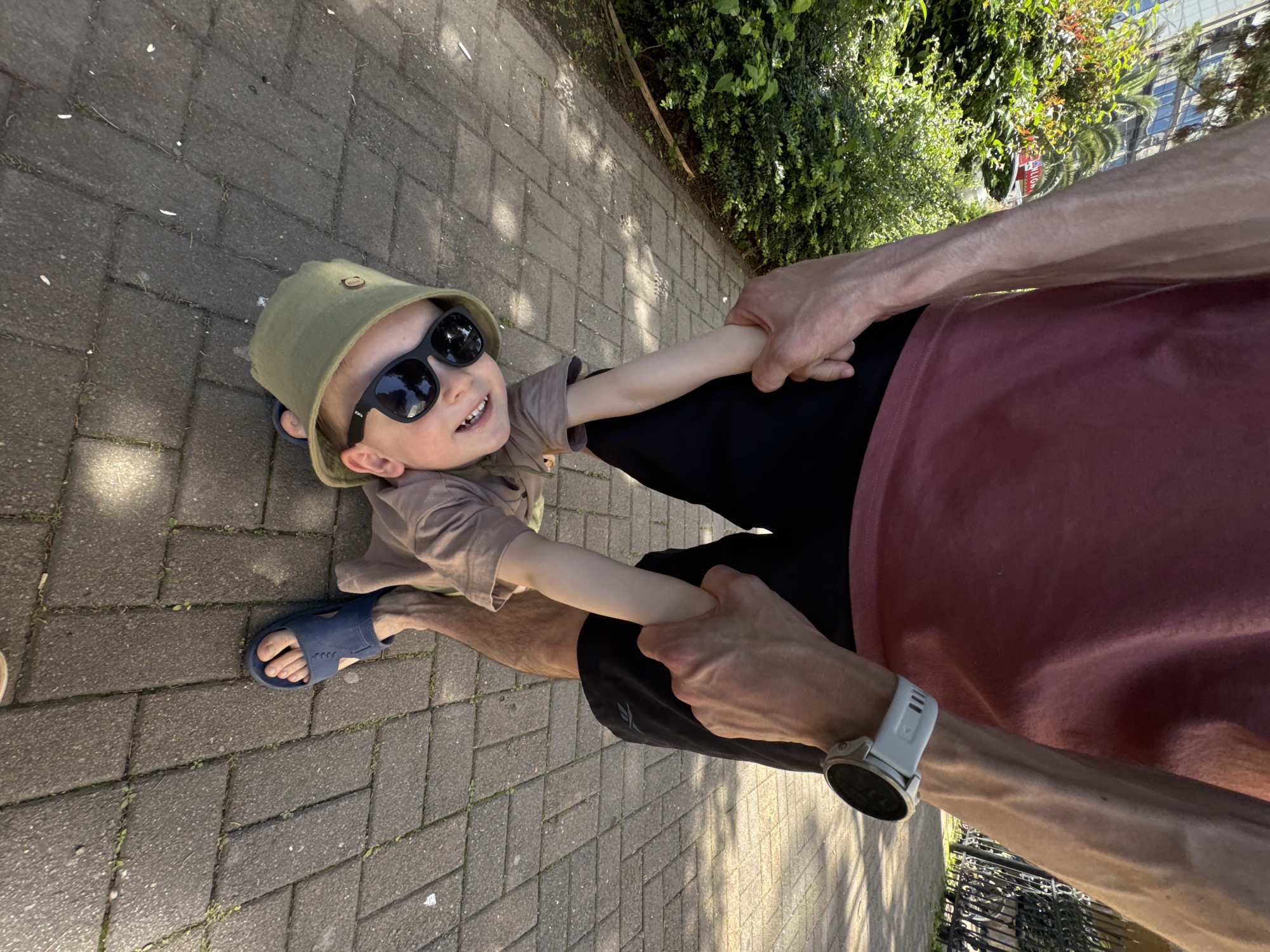 A low-angle shot shows a child with dark sunglasses and a bucket hat being held by a person wearing a maroon shirt and a smartwatch. The child is also wearing a brown shirt and blue sandals. They are standing on a brick-paved path next to greenery, possibly in Sochi, Russia.