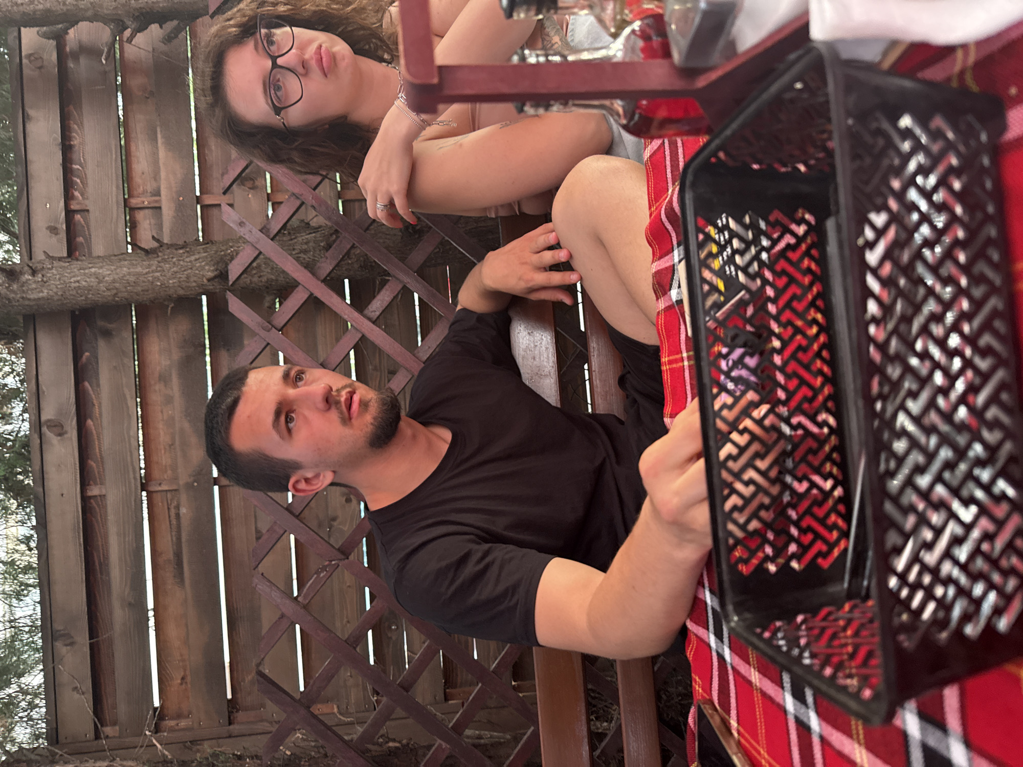 The image shows two young adults, a man and a woman, sitting at a table outdoors. The man, in the foreground, is wearing a black t-shirt and looking to his right. The woman, partially visible behind him, is wearing glasses and looking away. They are seated on a bench with a wooden lattice fence and trees in the background. The table is covered with a red and white plaid cloth, and a black basket is in the foreground. Bottles are visible on the right side of the table, suggesting a dining or drinking setting. The overall ambiance appears casual and relaxed, consistent with a restaurant or cafe in Sofia, Bulgaria.