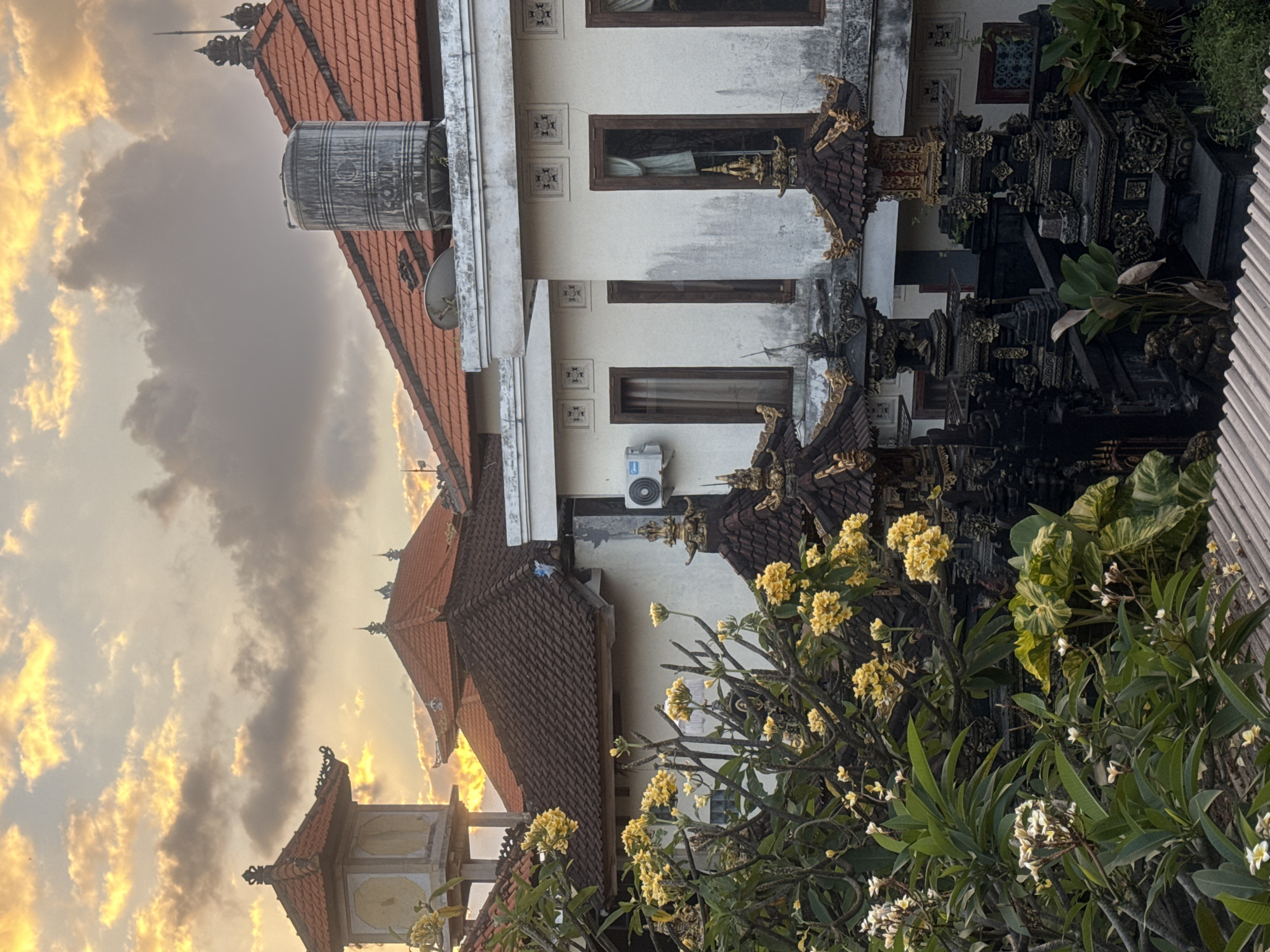 The image captures a serene view of buildings with traditional Balinese architecture in Kuta, Indonesia, at sunset. The sky is filled with dramatic clouds illuminated by the golden hues of the setting sun. In the foreground, a lush frangipani tree is in bloom, with its white and yellow flowers adding a touch of color to the scene.
The buildings feature terracotta tiled roofs, characteristic of Balinese design, some with intricate decorative elements. One prominent structure has a water tank on its roof. Below, a dark, ornate Balinese gate or shrine is visible, adding a cultural element to the urban landscape. The buildings appear to be residential or part of a complex, with windows and an air conditioning unit visible on one of the walls. The overall atmosphere is peaceful and picturesque, blending natural beauty with local architecture.