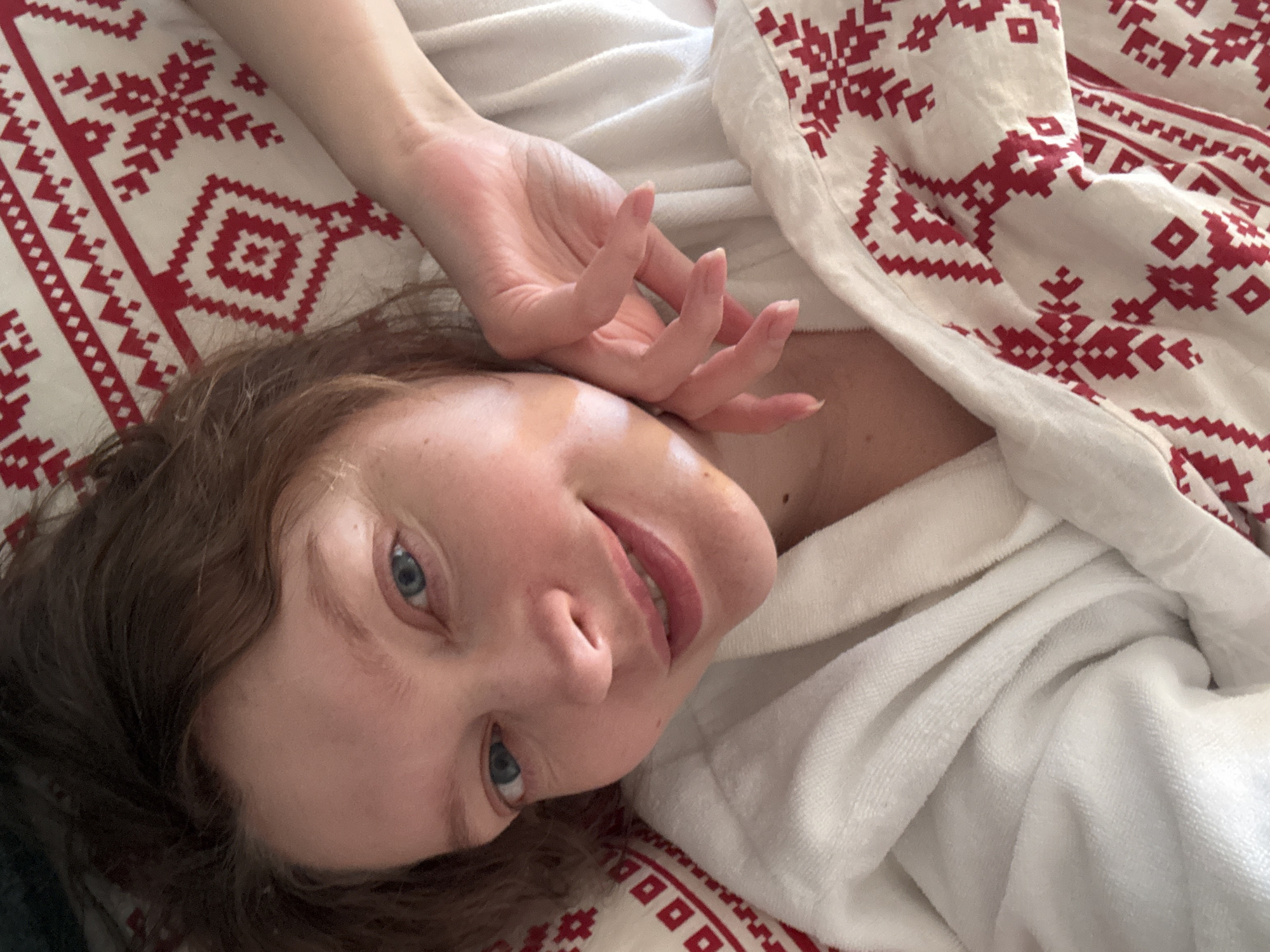 A woman with fair skin, brown hair, and blue eyes is lying down. Her head is tilted to the right, and she is smiling slightly. Her right hand is raised near her face. She is wearing a white robe and is lying on a white and red patterned fabric. The location is Unknown, Unknown.