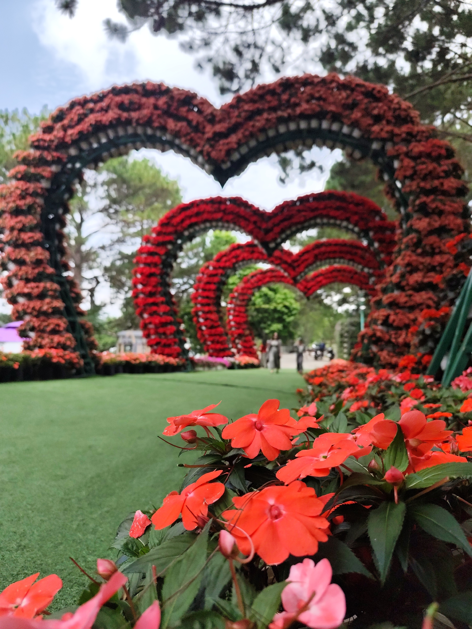The image shows a series of three heart-shaped arches decorated with red flowers, likely in a garden setting in Da Lat, Vietnam. The arches are positioned one behind the other, creating a tunnel-like effect leading into the distance. The foreground features a flower bed filled with red and some pink impatiens flowers, which are in sharp focus. Beyond the arches, two figures and a vehicle are discernible in the blurred background. The scene is set outdoors with a clear sky visible through the top of the arches, and the environment suggests a well-maintained public space or garden.