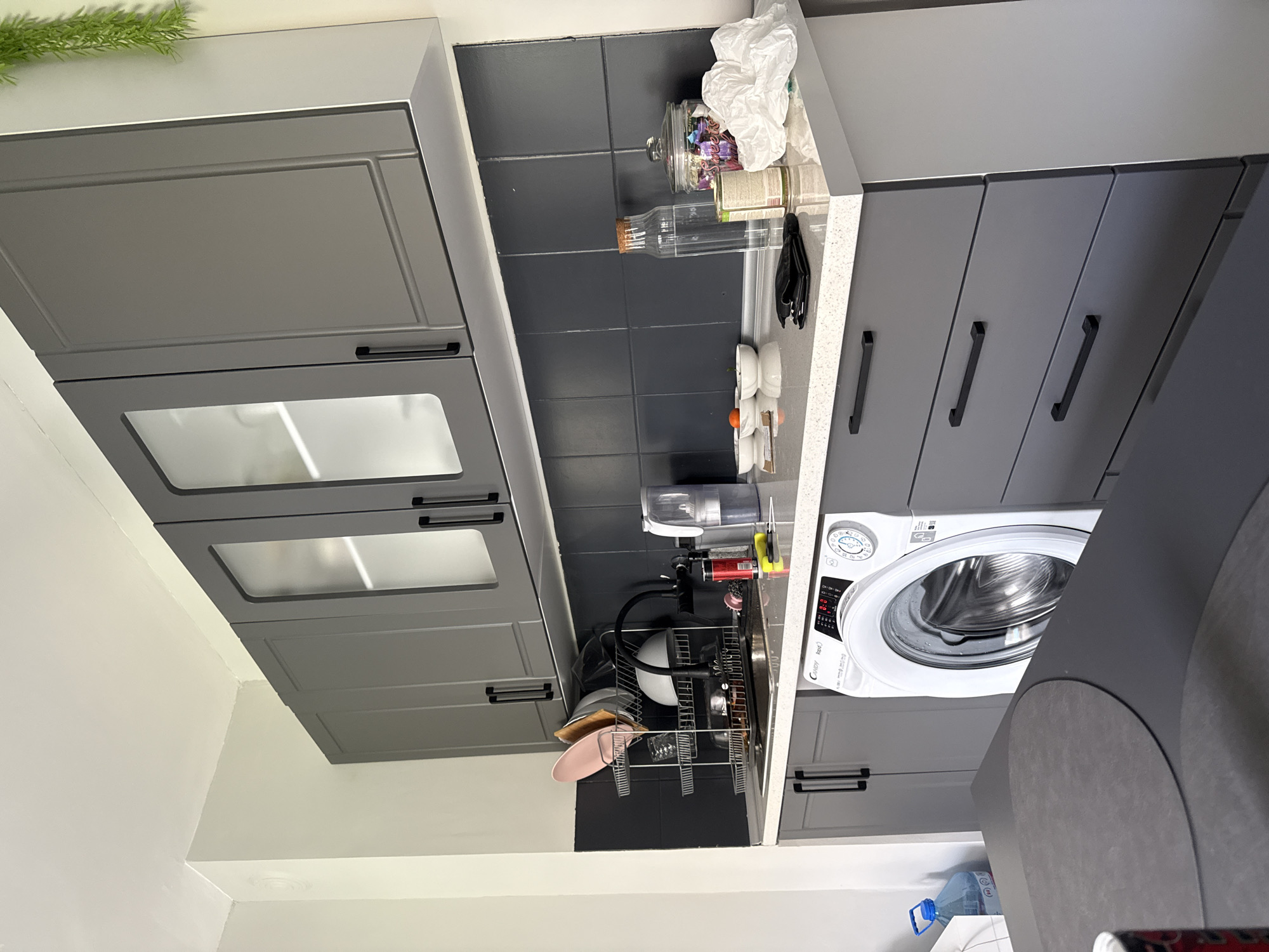 The image shows a kitchen interior. Gray cabinets are mounted on the wall, some with glass fronts. Dark gray tiles are on the wall between the upper and lower cabinets. On the counter, there are various items, including jars, bottles, dish rack with dishes, and a cleaning spray. A washing machine is positioned next to the counter. A water bottle is near the floor. The location is assumed to be in an indoor environment, potentially in the city of Stara Zagora, Bulgaria, based on the prompt's information request. No people or actions are visible in the image.