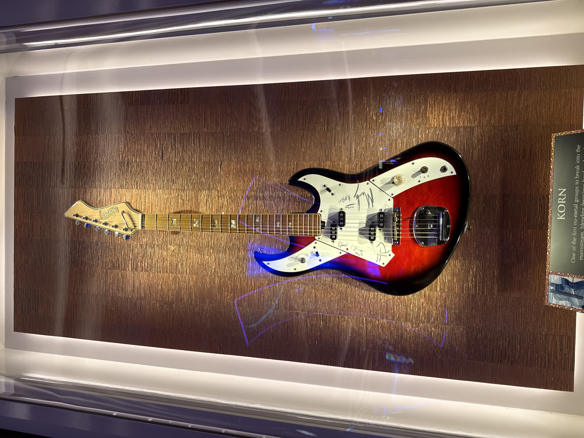 The image shows a signed electric guitar displayed in a glass case. The guitar has a red, black, and white body. It is mounted on a textured bronze-colored wall. There's a plaque to the right of the guitar that reads "KORN". The text describes Korn as one of the first nu-metal groups to break into the mainstream. The scene takes place indoors within a museum-like setting. The environment appears to be lit with recessed lighting at the top. The location of this scene is in the city of Hollywood Seminole Reservation, United States of America.