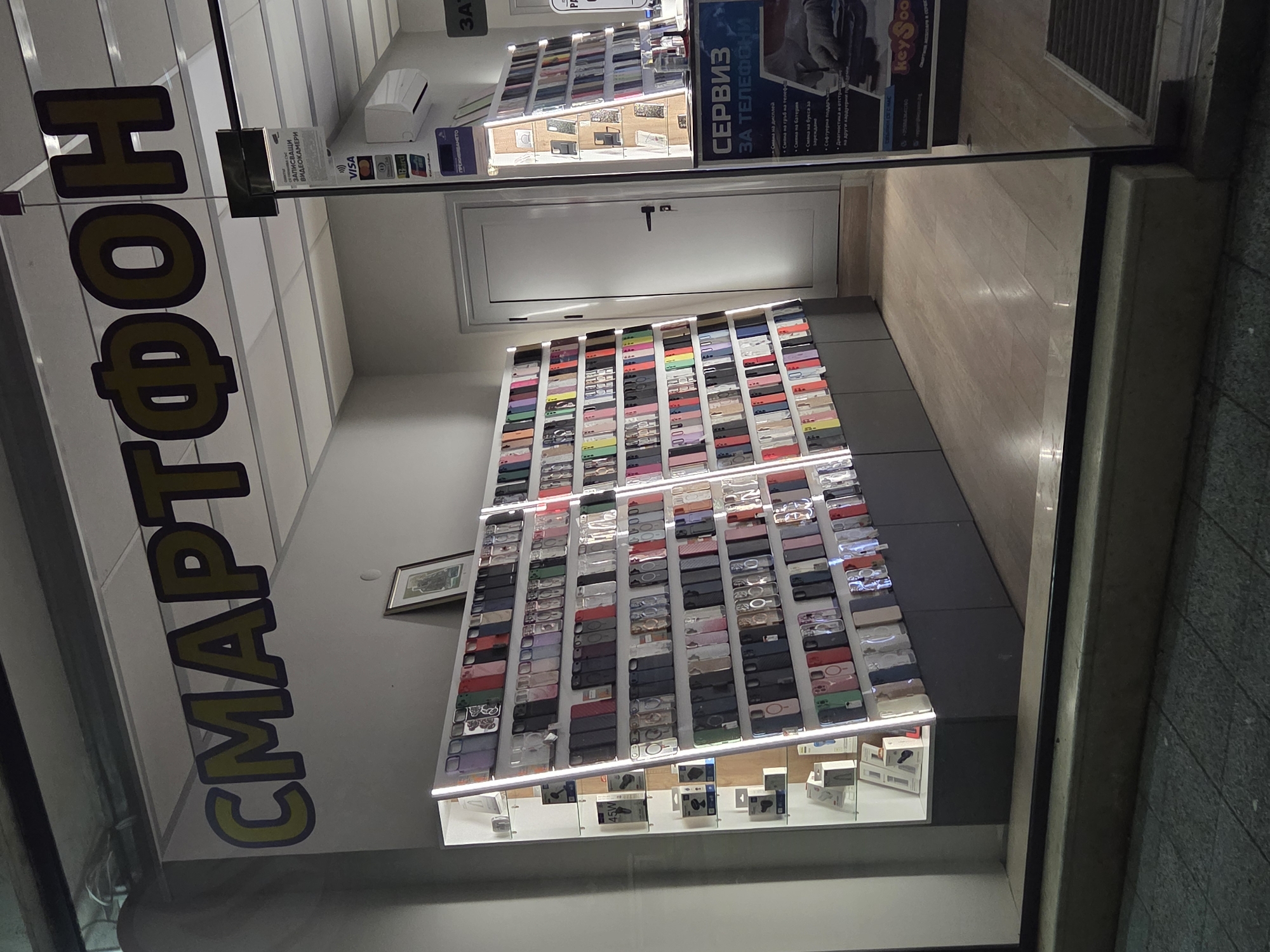 The image provides a view into a brightly lit mobile phone and electronics accessories shop in Pleven, Bulgaria, seen through a glass storefront. No people are visible.
The shop's interior features a large, dominant display unit against the back wall, showcasing a vast array of smartphone cases in various colors and designs, neatly arranged in grid-like rows. Below this extensive display, a lower glass shelf holds packaged electronic accessories such as chargers and headphones. To the left of the main display, a white, closed door is visible. Further back and to the right, an air conditioning unit is mounted on the wall above a smaller display of phone cases. The interior floor is composed of light-colored, wood-look planks, contrasting with the darker, textured exterior ground visible at the bottom right.
Multiple signs with Cyrillic text confirm the shop's offerings and services. A large sign prominently displays "СМАРТФОН" (SMARTPHONE), indicating the store's primary product. Other signs advertise "ЗАПИСВАЩИ ВИДЕОКАМЕРИ" (RECORDING VIDEO CAMERAS) and list accepted payment methods including "VISA", "Maestro", and "B-Card". A detailed service sign, titled "СЕРВИЗ ЗА ТЕЛЕФОНИ" (SERVICE FOR PHONES), enumerates services such as "Смяна на гръб" (Back cover replacement), "Батерия" (Battery replacement), "зарежда" (charging port repair), "Софтуерна" (Software service), "Диагнос" (Diagnosis), and "на други хардуери" (on other hardware). The brand name "KeySool" is also visible. The shop appears well-stocked and ready for customers, though the exact time of day or weather conditions are not discernible from the indoor lighting.