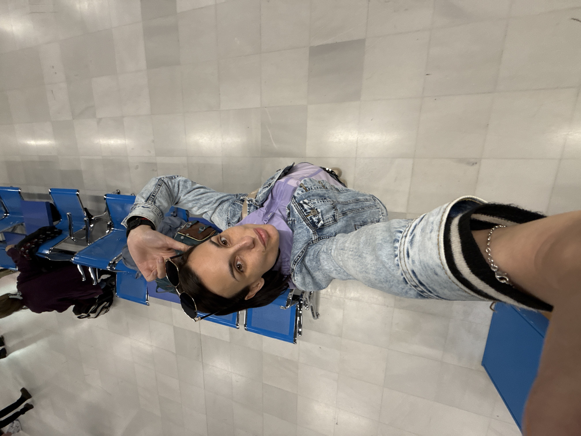 A person is lying on blue airport-style seating in what appears to be an airport or similar waiting area. The floor is light grey, composed of large square tiles. The person is wearing a light blue denim jacket over a purple top. The person is holding a pair of sunglasses. It is not possible to confirm the location of Kounoupidiana, Greece based on the provided image.
