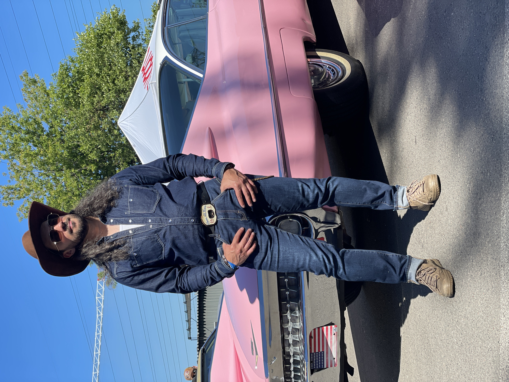 A man stands in front of a pink classic car, a tent in the background. He is wearing a brown cowboy hat, sunglasses, a denim shirt, and jeans. The man has long, dark, wavy hair and a beard. He is standing with his hands in his pockets. The pink car appears to be a vintage Cadillac. The scene is outdoors, with a bright blue sky and some trees visible. There is an American flag license plate on the front of the car. The environment is a paved surface, possibly a street or parking lot.
