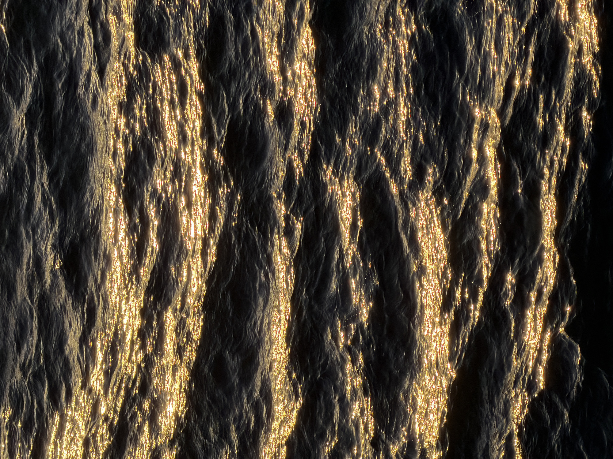 The image shows a close-up view of water reflecting sunlight. The water surface is rippled, creating varying levels of light and shadow. The sunlight is bright and golden, contrasting with the darker, shadowy areas of the water. The location is unknown.