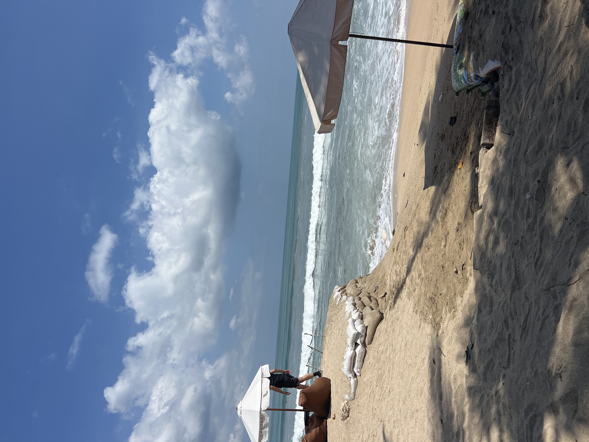 The image depicts a sandy beach scene in Kuta, Indonesia, under a bright blue sky with scattered white clouds. The ocean is visible in the background with moderate waves breaking along the shore. On the beach, two beach umbrellas are set up. One is white with the word "LAGOON" visible, positioned near a few large, brown beanbag chairs. A person wearing a black t-shirt and black shorts with a backpack is walking near this umbrella. The other umbrella is a lighter beige color and is closer to the foreground, partially obscuring the view of the ocean. The sand is light brown and appears slightly damp near the water. Along the edge of a small sandy embankment, there are sandbags reinforcing the shore. In the foreground, shadows from palm trees or other foliage are cast across the sand. The general atmosphere is that of a relaxed beach day.