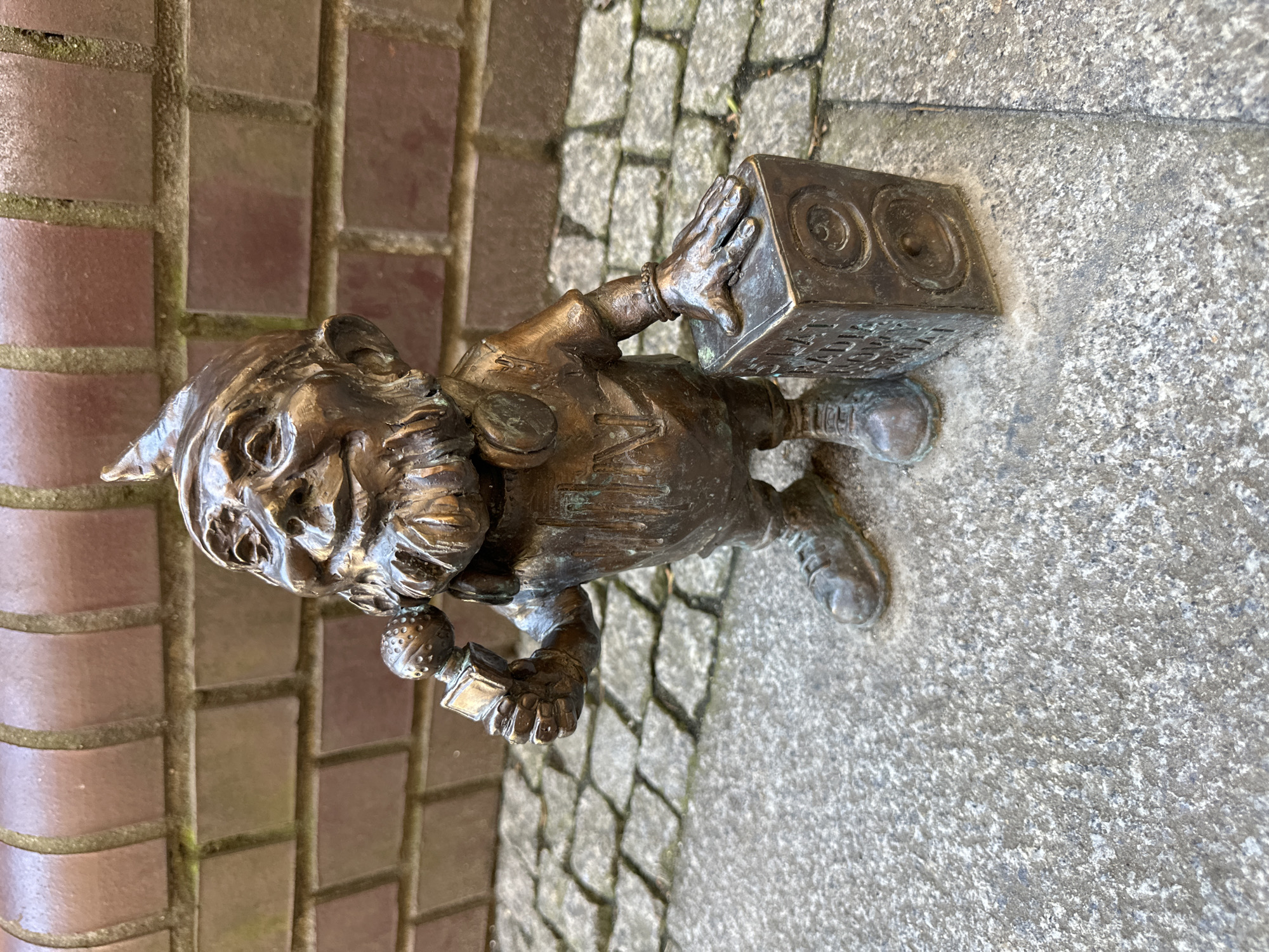 A bronze dwarf statue stands on a stone paved surface, partially adjacent to a brick wall. The dwarf, wearing a pointed hat, holds a boombox-shaped object in one hand and a microphone-like object in the other. The dwarf figure and the objects it holds are rendered in bronze. The statue is set in Wrocław, Poland, known for its gnome statues.