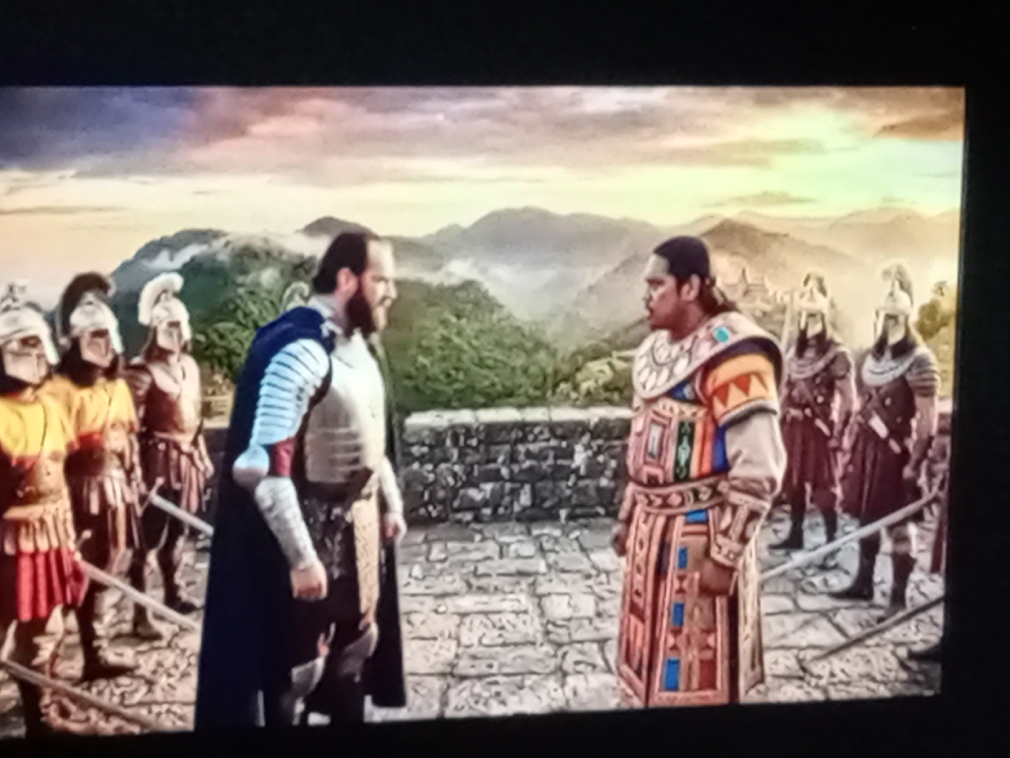 The image depicts an outdoor scene with two central figures engaged in conversation, standing on a paved courtyard before a stone wall. Behind the wall, a verdant, mountainous landscape with mist is visible under a cloudy sky. Several figures, dressed in armor and helmets, stand in formation to the left and right of the central pair. The central figures are a man with a beard, wearing a blue cape over silver armor, and another man in a richly decorated, multi-colored tunic. The man in the tunic appears to be speaking to the bearded man. The overall setting suggests a historical or fantasy context.