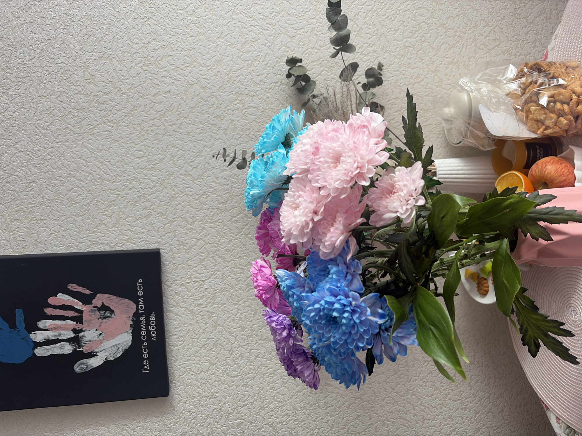 A close-up shot depicts a bouquet of vibrant flowers including blue, pink, and purple chrysanthemums, complemented by eucalyptus sprigs and greenery, in what appears to be a domestic setting. The flowers are positioned to the right, with a collection of items suggesting a casual, lived-in space, including a plastic container with crackers, fruit (apple and orange), and a juice bottle. To the left, a framed picture with handprints and text in Russian is visible against a textured wall. The text reads "Где есть семья, там есть любовь," translating to "Where there is family, there is love." Given the Russian text, the location is likely Saint Petersburg, Russia. There are no visible people or active interactions captured in the frame.