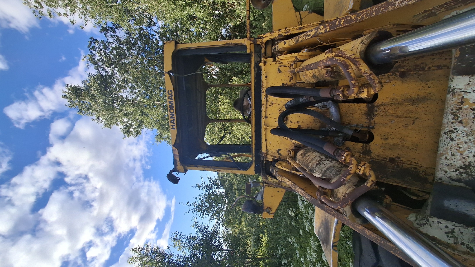 This media file shows a close-up, angled view of a yellow, weathered Hanomag loader. The loader is the primary object, with its cab and hydraulic components prominently featured. The machine appears old and somewhat rusty, suggesting it might be out of use or in need of repair.
Inside the cab, a person is visible, seated in the driver's position. Their face is partially obscured, but they appear to be looking forward. The steering wheel and dashboard are also visible.
The setting is outdoors, with lush green trees and foliage surrounding the loader. The sky is bright blue with scattered white clouds, indicating it is daytime. The presence of trees and foliage suggests a rural or semi-rural location, possibly a yard, field, or worksite. There are no specific architectural or geographical features that pinpoint Innolovo, Russia, beyond what is provided in the prompt.
The overall mood is neutral, focusing on the details of the machinery and the natural environment. No specific activity or event is depicted; it appears to be a still scene. The text "HANOMAG" is clearly visible on the side of the loader's cab.