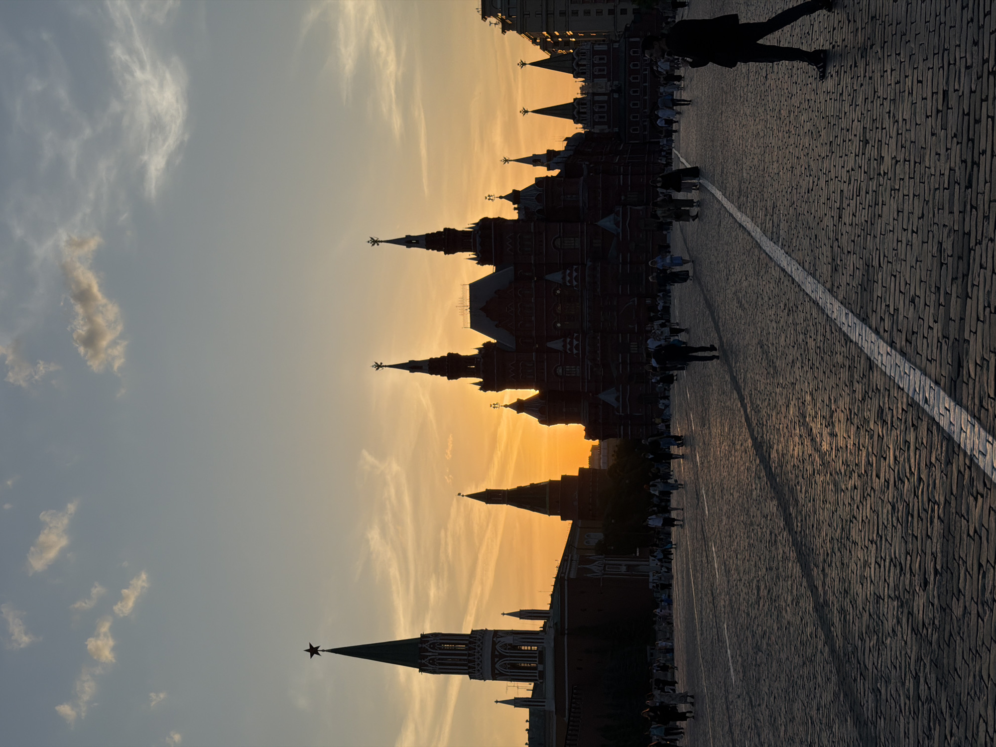 The image captures a sunset view of Red Square in Moscow, Russia. The silhouetted outlines of the Kremlin walls and towers dominate the skyline. Pedestrians, also silhouetted, walk along the cobblestone pavement. A white line marks a path on the square. The sky is streaked with clouds.