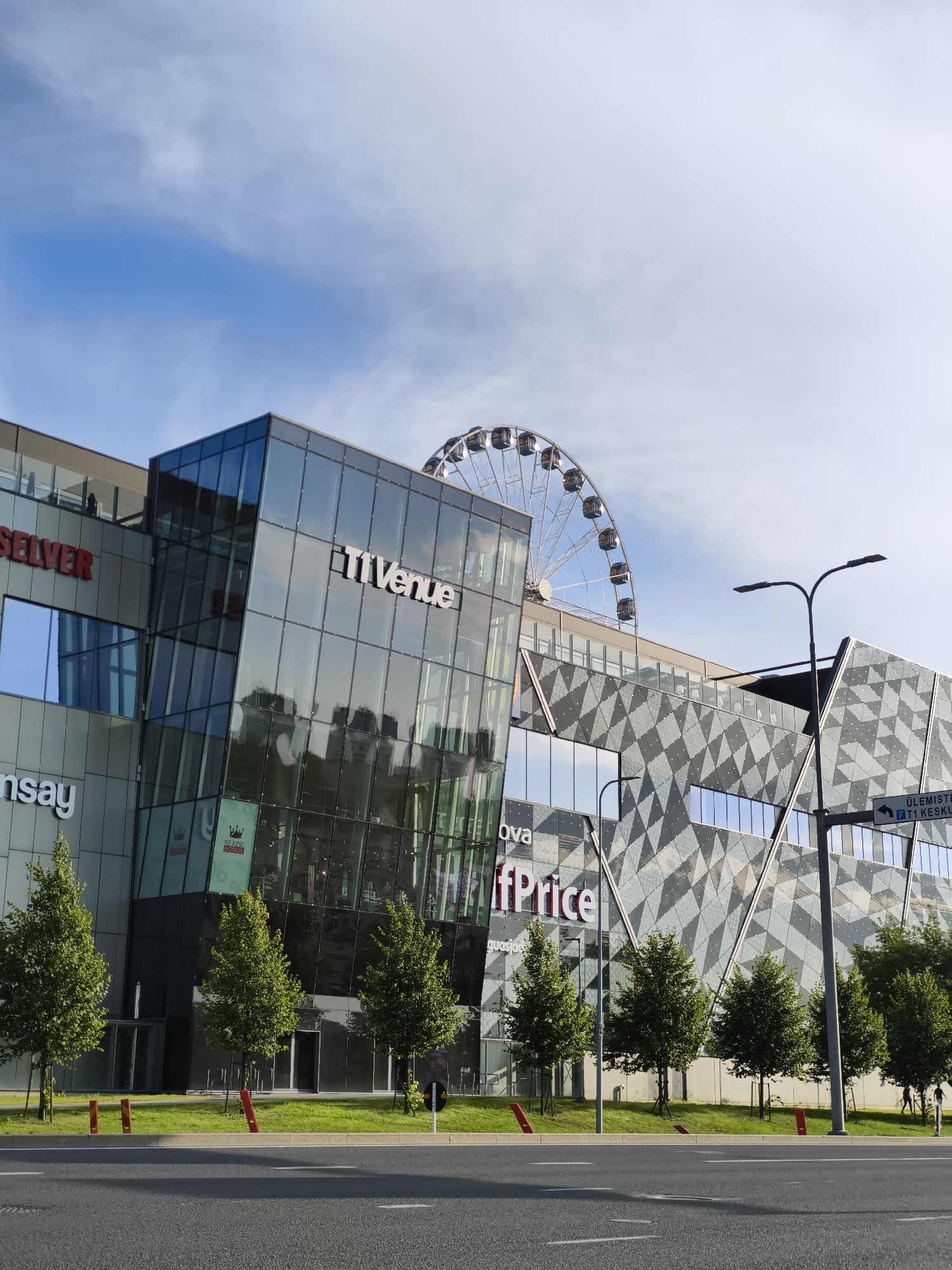 The image shows a modern shopping center with a large Ferris wheel visible in the background. The building features a lot of glass and geometric architectural elements. Signs indicate that it is the Ülemiste Keskus (Ülemiste Shopping Center), a prominent retail and entertainment complex located in Tallinn, Estonia, near the Tallinn Airport. There are trees and a street in the foreground. The sky is blue with some clouds.