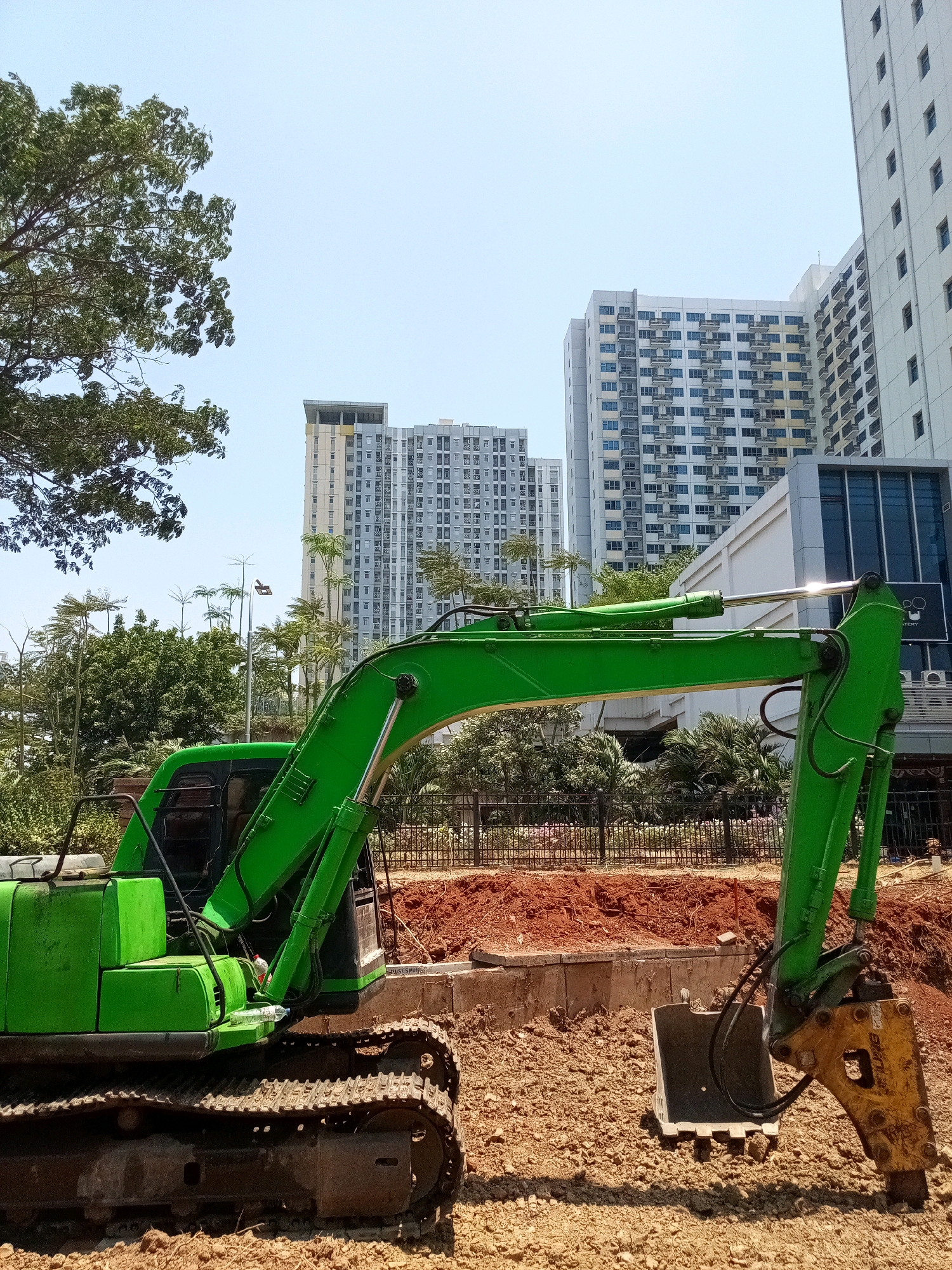 An excavator with a yellow jackhammer attachment is parked on a construction site in Bekasi, Indonesia. The excavator is bright green, with visible tracks and a cab. The ground is covered in dirt and small rocks. In the background, several tall residential buildings with numerous balconies are visible, suggesting an urban setting. Trees and streetlights are also present in the background, along with a black metal fence. The sky is clear and blue.