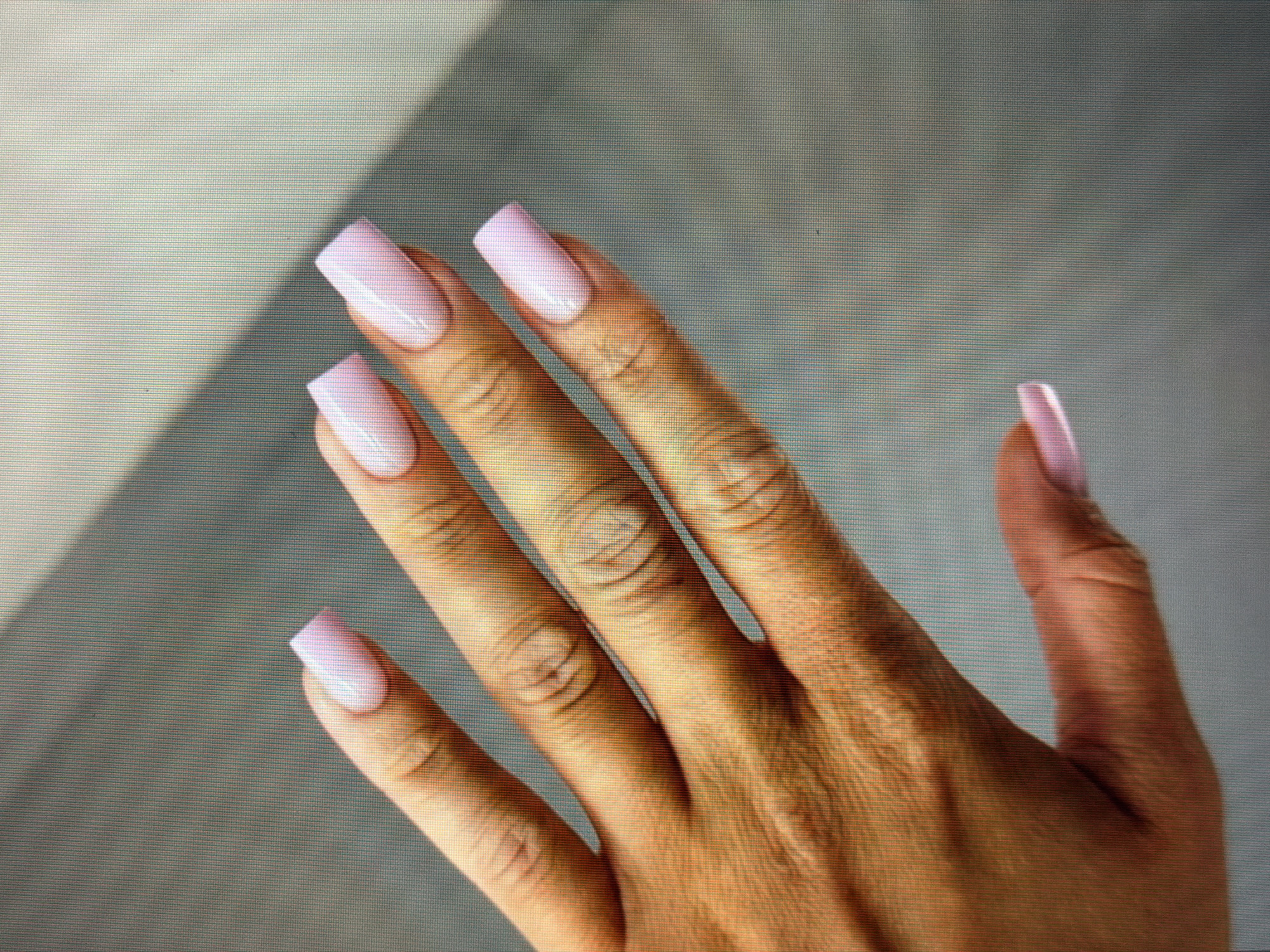 A close-up, eye-level shot shows the manicured hand of a person with light pink, square-shaped nails. The hand is positioned diagonally from the lower left to the upper right of the frame. The background is a softly blurred, neutral gray wall with a diagonal shadow cast across it from the upper left. The lighting is soft and diffused. The image does not depict a specific location in Sirius, Russia.