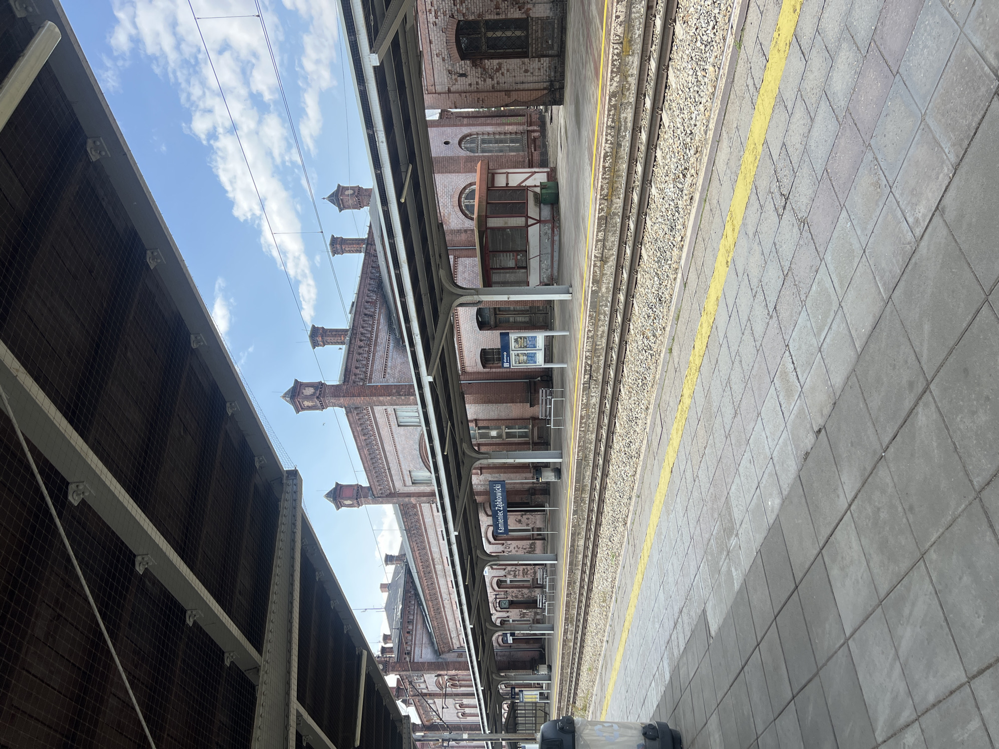This is a photograph of a train station platform in Kamieniec Ząbkowicki, Poland. The scene is outdoors, under a bright blue sky with scattered white clouds, suggesting daytime with pleasant weather. The platform itself is paved with gray tiles and bordered by a yellow line. Adjacent to the platform are train tracks with gravel ballast.
To the right of the tracks, an old brick building with arched windows and ornate brickwork serves as the station structure. This building is covered in part by a covered walkway supported by arched metal columns, typical of older European train stations. Chimney-like structures with pointed roofs rise from the building's top.
A blue sign is visible on a pole, mounted under the covered walkway, bearing white text. Although slightly blurry, the text appears to read "Kamieniec Ząbkowicki". There are no people clearly visible in the frame. The overall impression is of a historic, functional train station operating on a bright, clear day. The perspective is from the platform, looking along the tracks and towards the station building. A portion of a dark, covered area, possibly another platform or part of the station's roof structure, is visible in the foreground on the left.