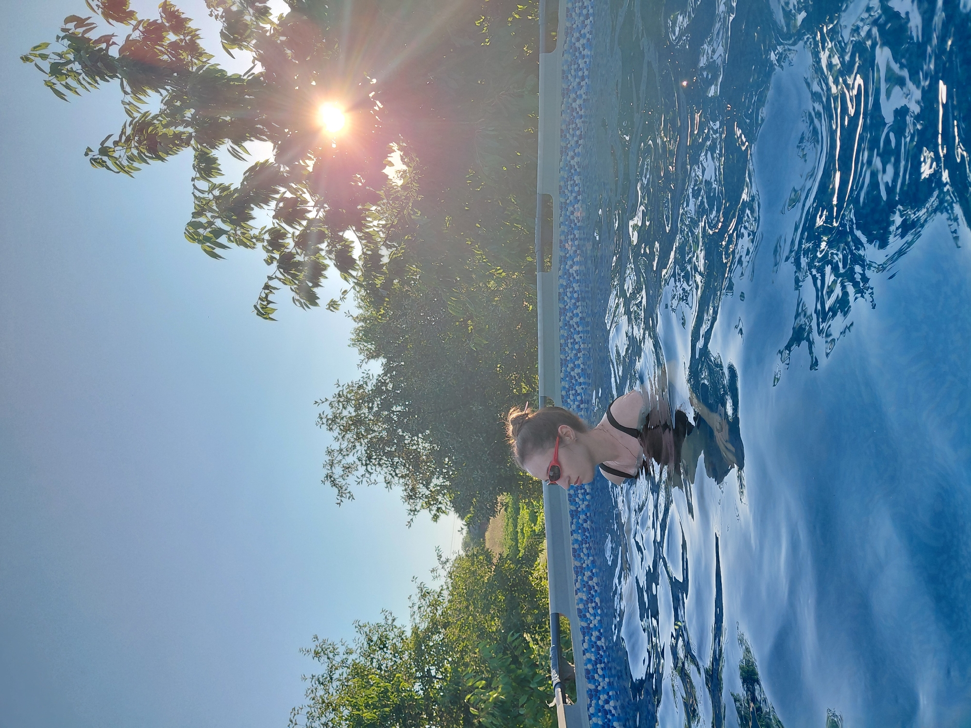 In a shallow, above-ground swimming pool with a patterned blue and white tiled border, a young woman with her hair pulled up in a bun is swimming. She is wearing red swimming goggles and a black swimsuit. The pool water is a vibrant blue and is rippling. The background shows a sunny day with green trees and foliage, and the sun's rays are shining brightly through the leaves of a tree on the right side of the image. The scene appears to be in a backyard or outdoor area.