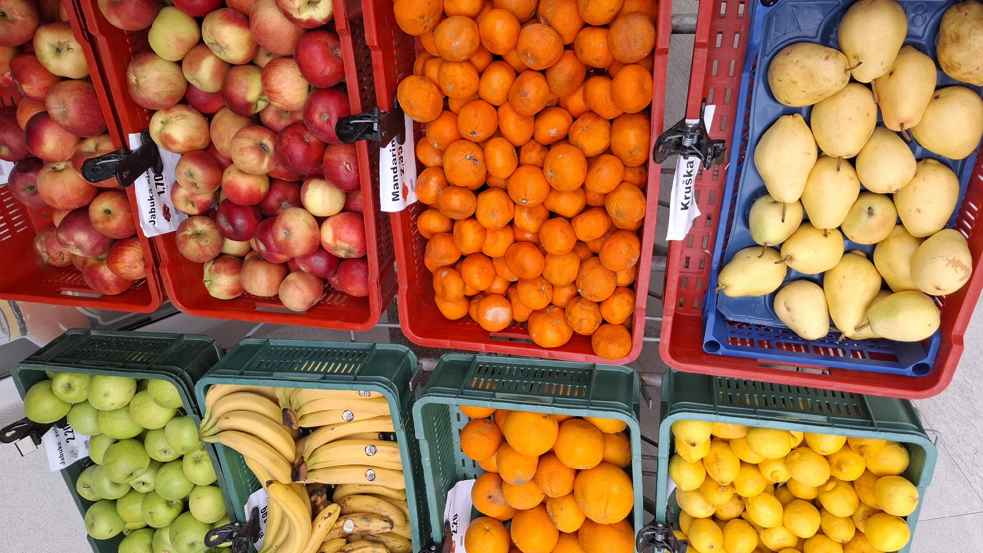 The image displays an outdoor fruit stand or market stall located in Boreti, Montenegro. Six plastic crates, arranged in two vertical columns, are filled with various fresh fruits.
On the left column, from top to bottom:
1. A dark green plastic crate contains numerous green apples. A white paper label, secured by a black clip, displays "Jabuke zelene 2,20", indicating green apples priced at 2.20.
2. Below this, another dark green plastic crate holds multiple bunches of yellow bananas. A partial white label showing "1,00" is visible.
3. At the bottom, a third dark green plastic crate is filled with bright yellow lemons.
On the right column, from top to bottom:
1. A red plastic crate contains a mixture of red and yellow apples. A white paper label with a black clip reads "Jabuka 1,70", indicating apples priced at 1.70.
2. Below this, a second red plastic crate holds a large quantity of small, vibrant orange mandarins. A white paper label states "Mandarina 2,00", indicating mandarins priced at 2.00.
3. At the bottom, a third red plastic crate contains a blue plastic tray filled with light yellow pears. A white paper label with a black clip indicates "Kruška", which is Croatian/Serbian for "Pear".
The crates are positioned on a light-colored, possibly tiled or concrete, ground surface. A light-toned wall or background structure is partially visible in the upper left. No individuals, actions, or interactions are depicted within the frame.