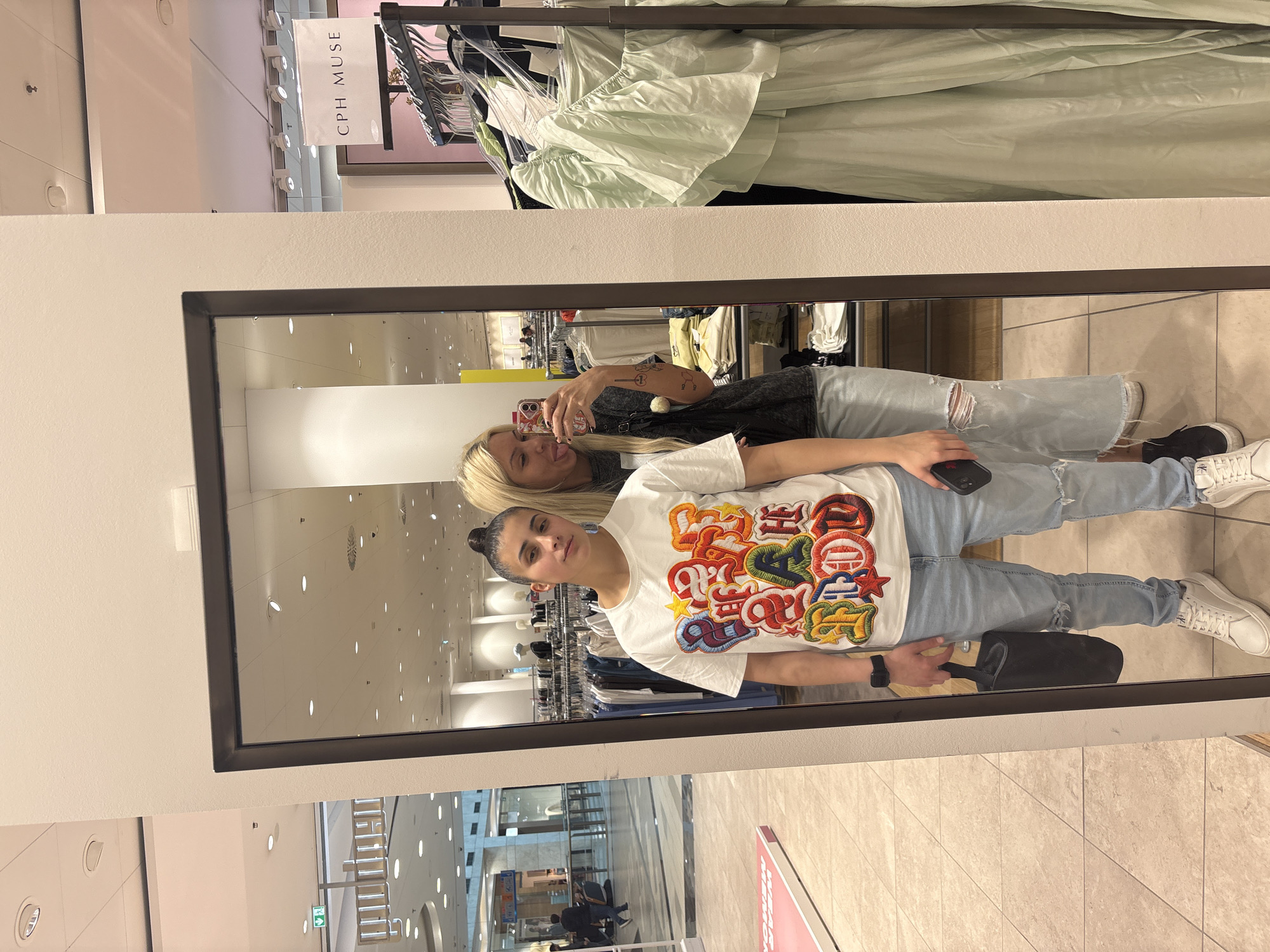 The image captures a mirror selfie of two women taken inside what appears to be a modern shopping mall or large department store in Sofia, Bulgaria.
The central feature is a large, dark-framed mirror reflecting the two subjects and the surrounding retail environment. In the reflection, the woman closer to the mirror has blonde hair, wears a dark t-shirt, ripped light-wash jeans, and black sneakers. She is holding a smartphone with a colorful case, extending her arm to take the photo, and has her tongue playfully sticking out. Visible tattoos adorn her right arm. Behind her, the second woman has dark hair styled in a bun. She is wearing a white t-shirt with a distinctive, multi-colored graphic design featuring stylized letters (parts of which appear to spell "COLOR"), light-wash ripped jeans, and white sneakers. She looks directly towards the camera with a neutral expression, holding a black rectangular object and a black smartphone.
The background outside the mirror, above the reflection, shows clothing racks with light green garments, along with a white hanging sign displaying the text "CPH MUSE". Through the mirror's reflection, the interior of the mall is visible, including bright overhead lighting, tiled floors, glass railings, and other distant storefronts. The scene conveys a casual shopping excursion during daytime.