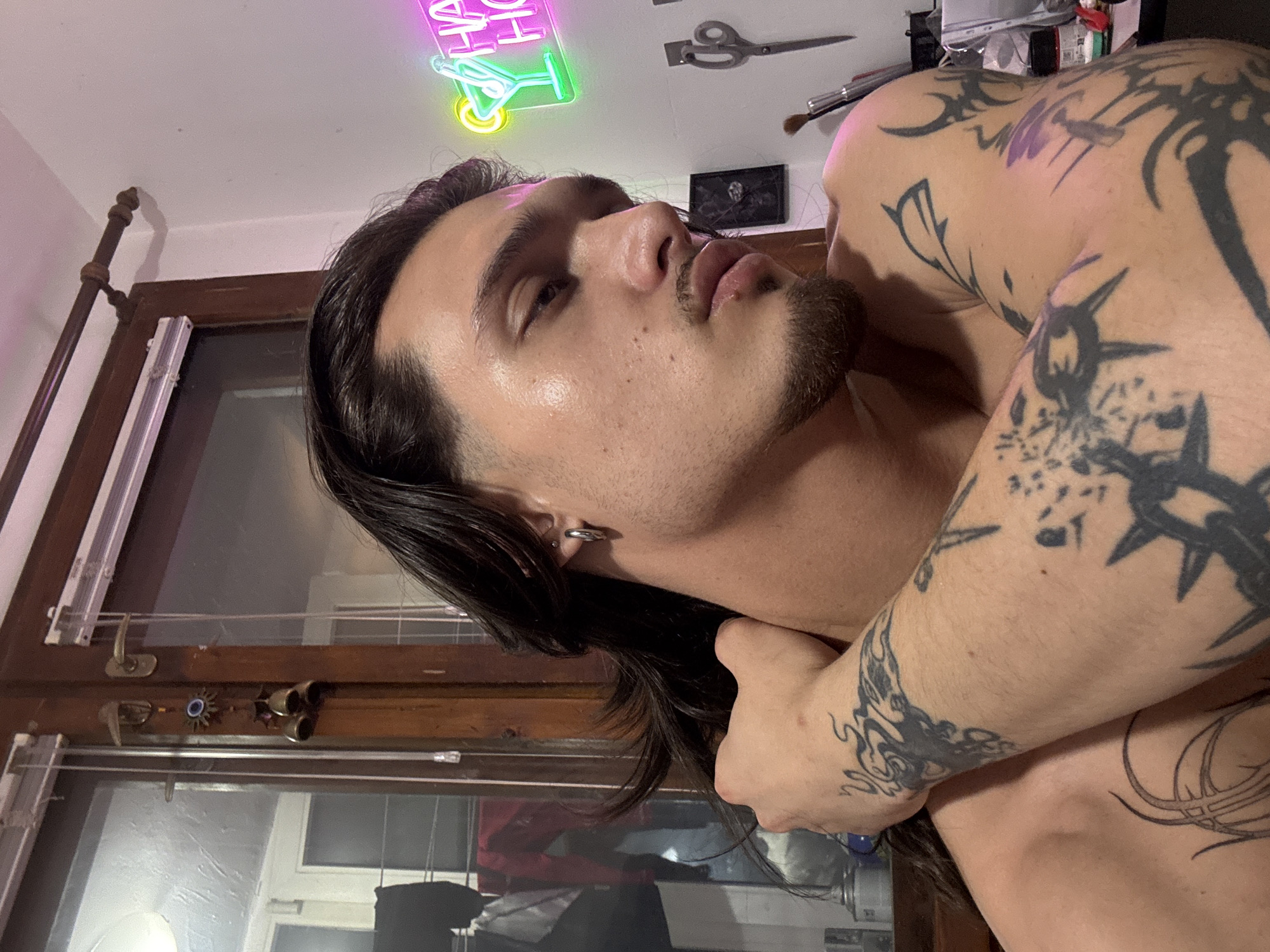 A young man with long dark hair, a stubble beard, and prominent tattoos on his arms and shoulder is depicted indoors, likely in a bedroom. He is shirtless, seen from the chest up, and appears to be lying down or leaning back, looking upwards and slightly away from the camera. His right arm is bent, with his hand resting near his neck. He wears a silver hoop earring in his left ear.
The background reveals a room with a relaxed, somewhat eclectic ambiance. To the left, a dark wooden window frame with white blinds or shutters is visible, alongside what appears to be part of a bed frame or decorative pole. To the right, a white wall features a neon sign that partially reads "HAPPY HOUR," with "HA" and "HO" clearly visible, along with the outline of a cocktail glass glowing in pink/purple and green/yellow. Below the sign, a pair of scissors hangs on the wall, and various items like brushes and containers are arranged on a surface.
The man's skin shows some texture and minor blemishes. His tattoos are extensive, featuring black ink tribal or abstract patterns, including what resembles a barbed wire or crown of thorns design on his right upper arm. His expression is neutral and calm, with slightly parted lips. The lighting in the room is dim, with the neon sign providing a significant light source, suggesting it might be evening or night. The image captures a personal moment, possibly a relaxed pose or selfie. No specific visual cues within the image confirm the location as Sofia, Bulgaria, beyond the provided context.