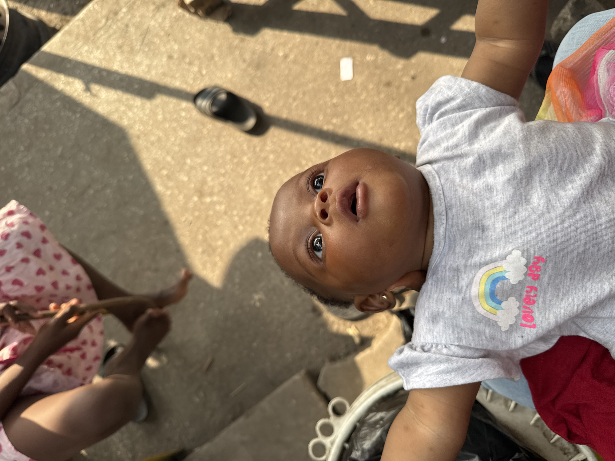 An overhead shot captures a Black baby lying down, looking upwards. The baby is wearing a grey t-shirt with a rainbow and the text "lovely day" printed on it. The baby's arms are outstretched, and its head is slightly tilted. In the background, the legs of another person, likely an adult, are visible. This person is wearing a pink dress with a heart pattern. The environment appears to be outdoors on a concrete surface, possibly a street or sidewalk, with a shadow cast by what seems to be a fence or railing. The lighting suggests it is daytime, with sunlight creating strong contrasts. The scene is situated in Badagry, Nigeria.