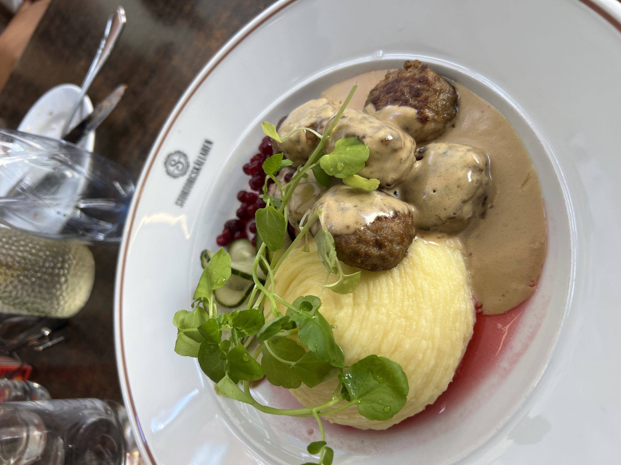 A white plate with a brown rim sits on a wooden table. On the plate is a serving of Swedish meatballs in a creamy sauce, accompanied by a quenelle of mashed potatoes, a garnish of watercress, sliced cucumbers, and lingonberries. A logo with the letter "S" inside a circular design and the text "STORRÄSKULLAREN" is visible on the rim of the plate. In the background, a blurred glass of water and a bottle of sparkling water are present. The image appears to be taken in a restaurant in Stockholm, Sweden.