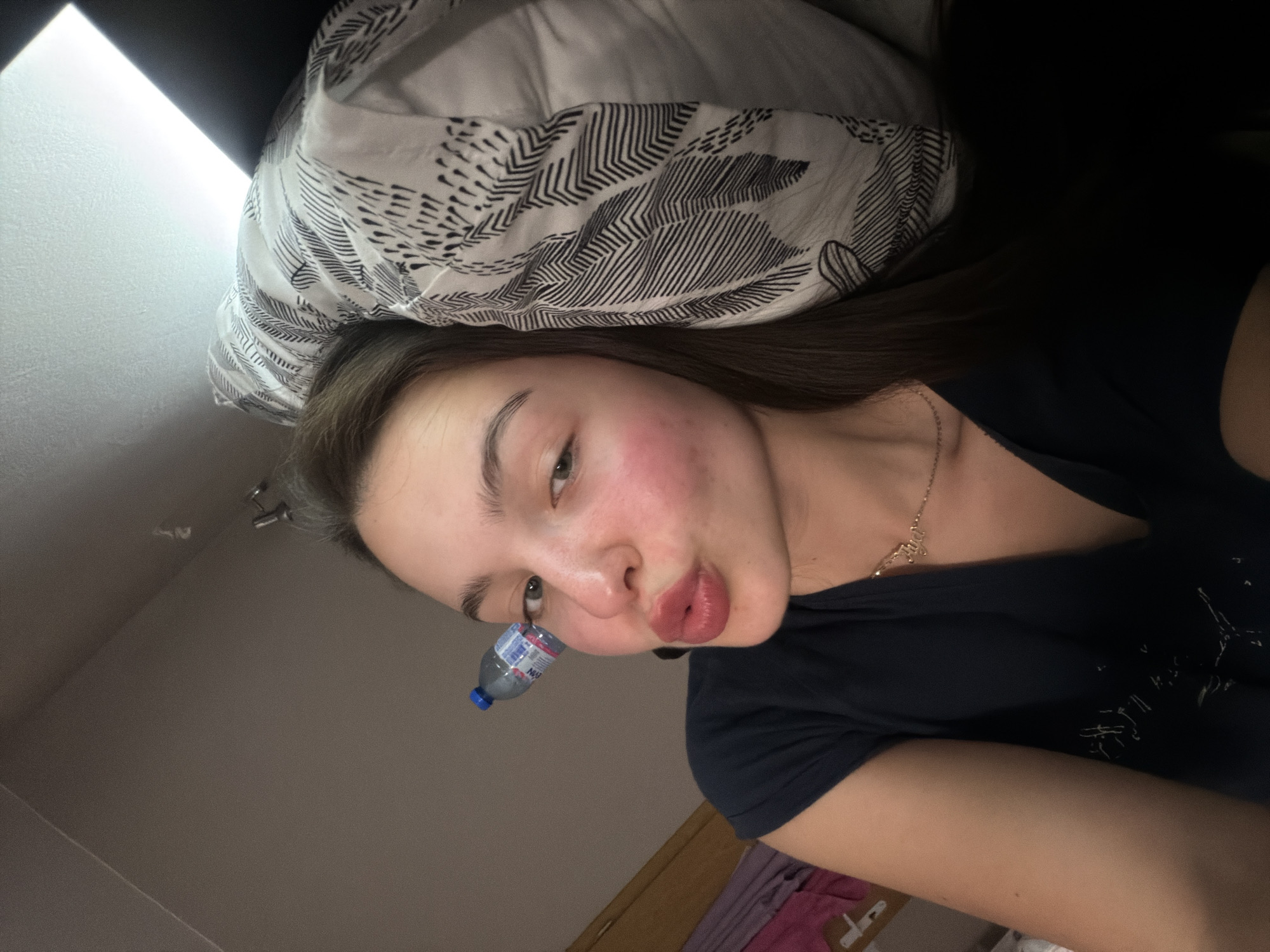 A young woman is photographed in an indoor setting, likely a bedroom, given the pillow behind her head. She has fair skin, dark hair, and is wearing a dark-colored t-shirt. A small water bottle is positioned near her face. A glimpse of furniture is visible in the background. The location within the city of Unknown, Unknown is unspecified and cannot be determined from the image.