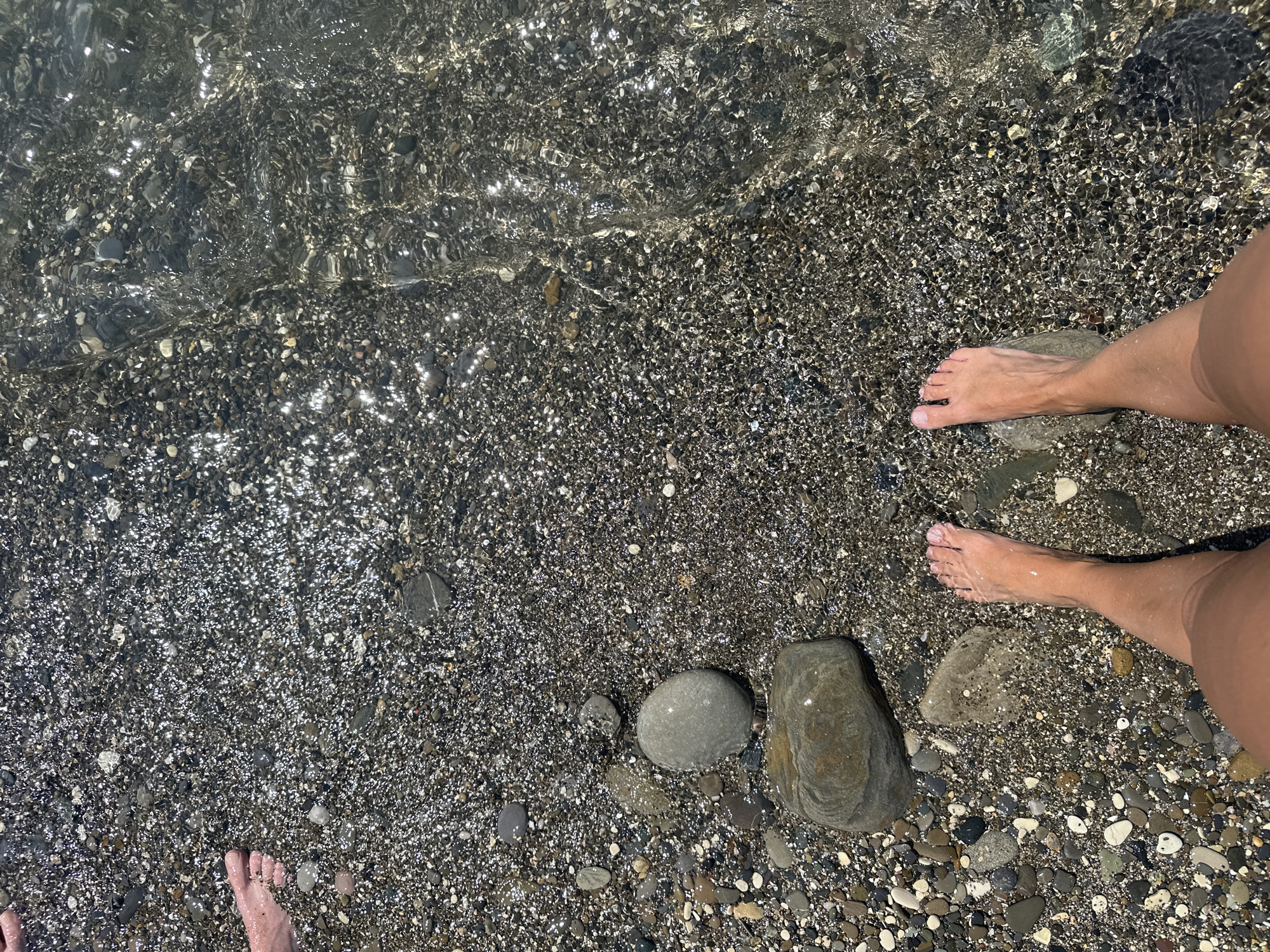 The image shows a person's bare feet standing on a pebble beach with shallow, clear water flowing over it. The pebbles are dark gray and black, with some lighter colored stones interspersed. The water is reflecting the sunlight, creating a shimmering effect. In the background, another person's feet are visible on the left side of the frame, partially submerged in the water. The scene is likely a beach in Sochi, Russia, known for its pebble beaches and clear waters. The focus is on the textures of the pebbles and the water, with the feet providing a sense of scale and human presence.
