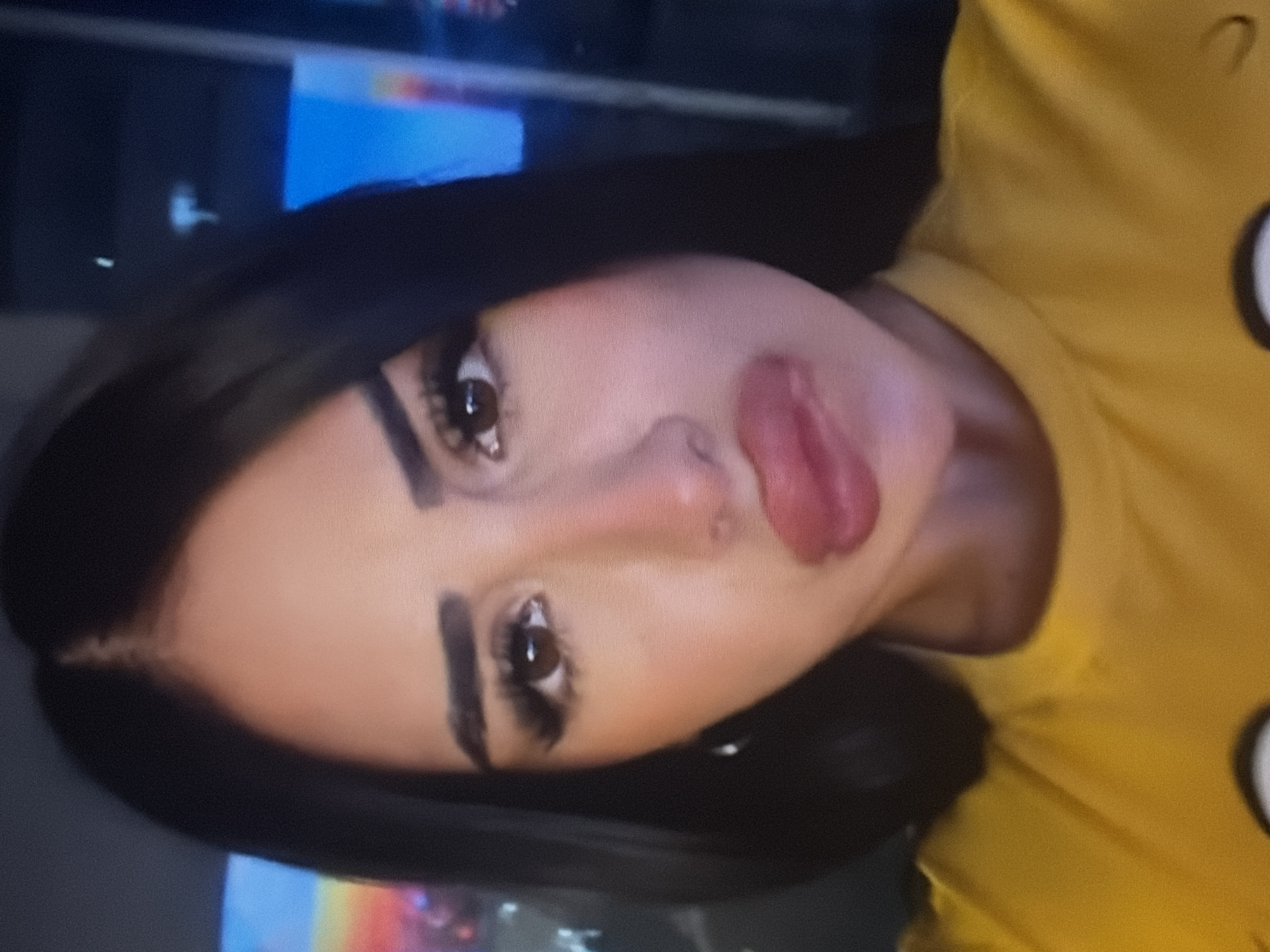 The media file is a close-up, vertically oriented, low-resolution photograph of a young person's face, appearing to be a selfie. The subject has dark hair, prominent eyebrows, and is wearing makeup that includes dark eyeliner and pink lipstick. Their lips are slightly parted, and their tongue is visible, giving a playful or provocative expression. The subject is wearing a bright yellow garment, possibly a t-shirt, with a dark outline of what appears to be a character's face on it, visible on the right side of the frame.
The background is dark and out of focus, with some blurred colorful lights suggesting an indoor setting, possibly a room with screens or a television on. There is no clear indication of the time of day or weather. The overall mood appears to be casual and possibly attention-seeking due to the pose and makeup. No text is visible in the image.