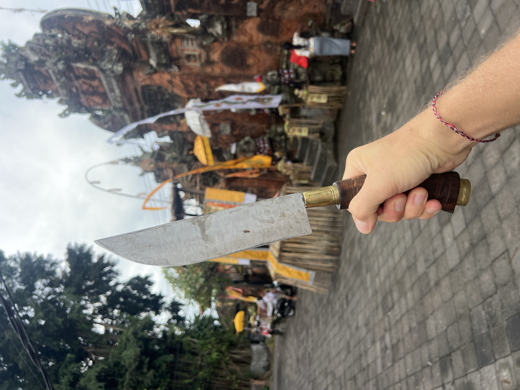 A person's hand holds a machete in front of a temple in Sukawati, Indonesia. The machete has a worn, silver-colored blade and a brown wooden handle with brass accents. The person is wearing a red and white bracelet. In the background, a large, ornate temple structure is visible, adorned with decorations and flags. People can be seen moving around the temple grounds. A powerline crosses over the tree. The ground is paved with gray bricks.