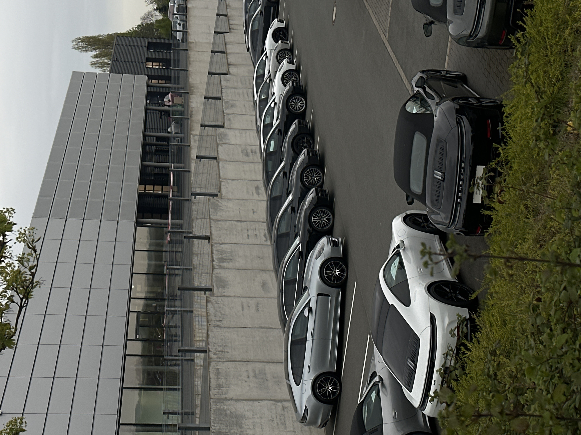 The image depicts a parking lot in Holzwickede, Germany, adjacent to a modern, multi-story building with a gray tile facade and large windows. A row of parked cars, primarily black and silver Porsches, occupies the parking spaces. The cars are lined up along a concrete wall with metal grating. The sky is overcast. Foliage is visible on the right side of the frame.
