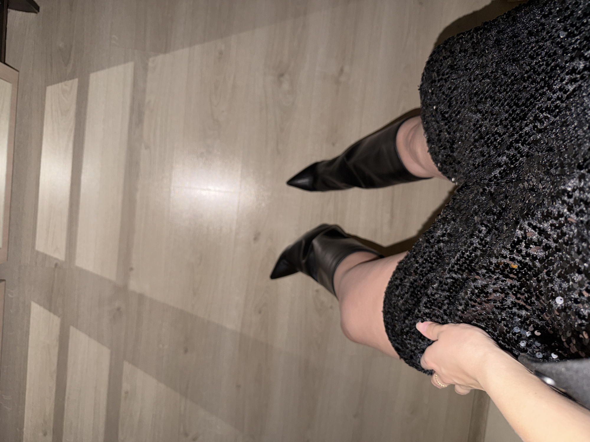 A floor-level shot from above depicts a person wearing a black sequined dress and black leather boots. The person's hand is holding the hem of the dress. The floor appears to be made of light wood, with shadows cast across it. The location is not identifiable as being in Izhevsk, Russia.
