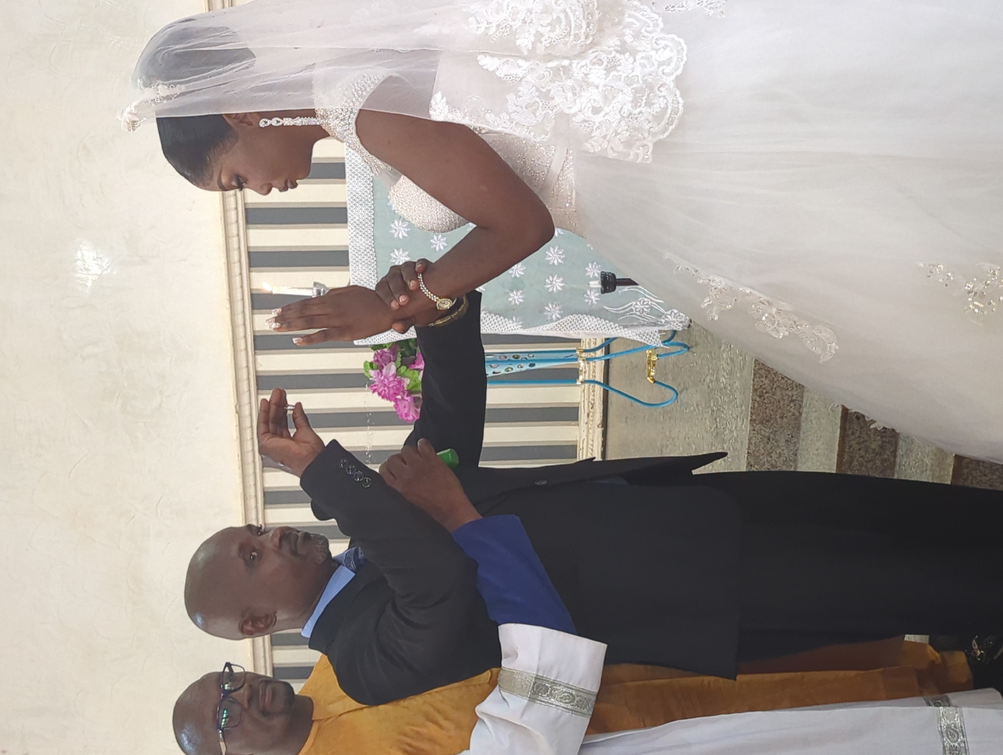 The image captures a wedding ceremony in Narayi, Nigeria. The foreground is dominated by a bride in a white, embellished wedding gown and veil. Her left hand is raised, and a man in a dark suit is holding her right wrist. This man, who appears to be officiating or involved in the ceremony, is also holding a ring in his left hand and a green object, possibly a small bottle or container, in his right hand. To the left of the officiant is another man in a yellow robe, and behind him is a partially visible man with glasses. The background features a striped wall and a decorated altar with flowers. The overall setting suggests an indoor event, likely a church or a reception hall.