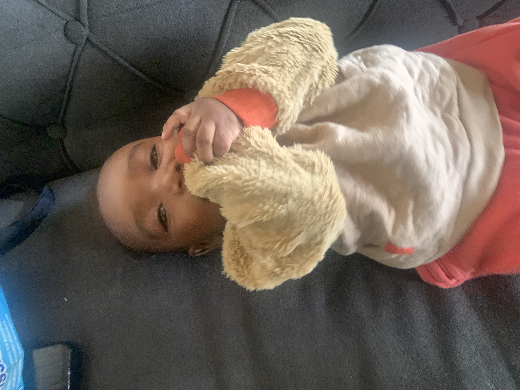 An infant is lying on a gray upholstered sofa. The infant is wearing a beige fuzzy jacket with orange cuffs over a white top and pink pants. The infant is holding their hand to their mouth. A portion of a blue package is visible at the left edge of the frame. There is no evidence to suggest the location is Barakin Akawo, Nigeria.