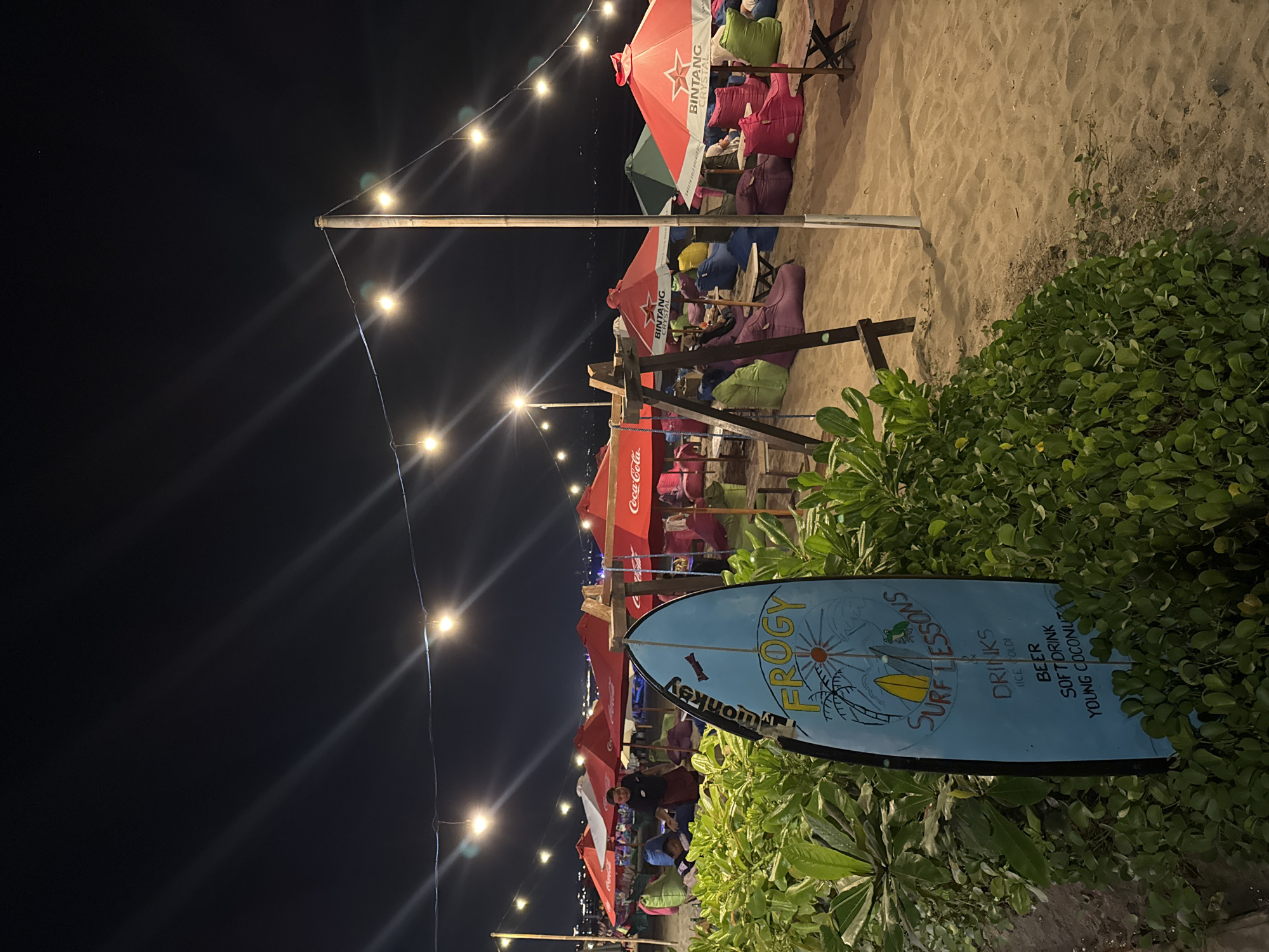 A night-time beach scene in Kuta, Indonesia, is depicted, characterized by soft sand, dimly lit open-air seating arrangements, and decorative string lights overhead. In the foreground, a light blue surfboard-shaped sign, angled towards the viewer, advertises "FROGY SURF LESSONS" and lists "DRINKS (ICE COLD)", "BEER", "SOFT DRINK", and "YOUNG COCONUT". The sign is surrounded by lush green foliage.
Further back on the sandy beach, numerous red and green umbrellas are open, casting shadows on the ground and sheltering beanbag chairs in various colors, including pink, purple, and green. Small wooden tables are scattered among the seating. A wooden swing set is visible to the left of the umbrellas. In the midground, a person wearing a dark shirt and apron is interacting with another person sitting at one of the tables. The background shows the ocean and the distant lights of the city. The overall atmosphere is relaxed and inviting, indicative of a beachside establishment open for evening service.