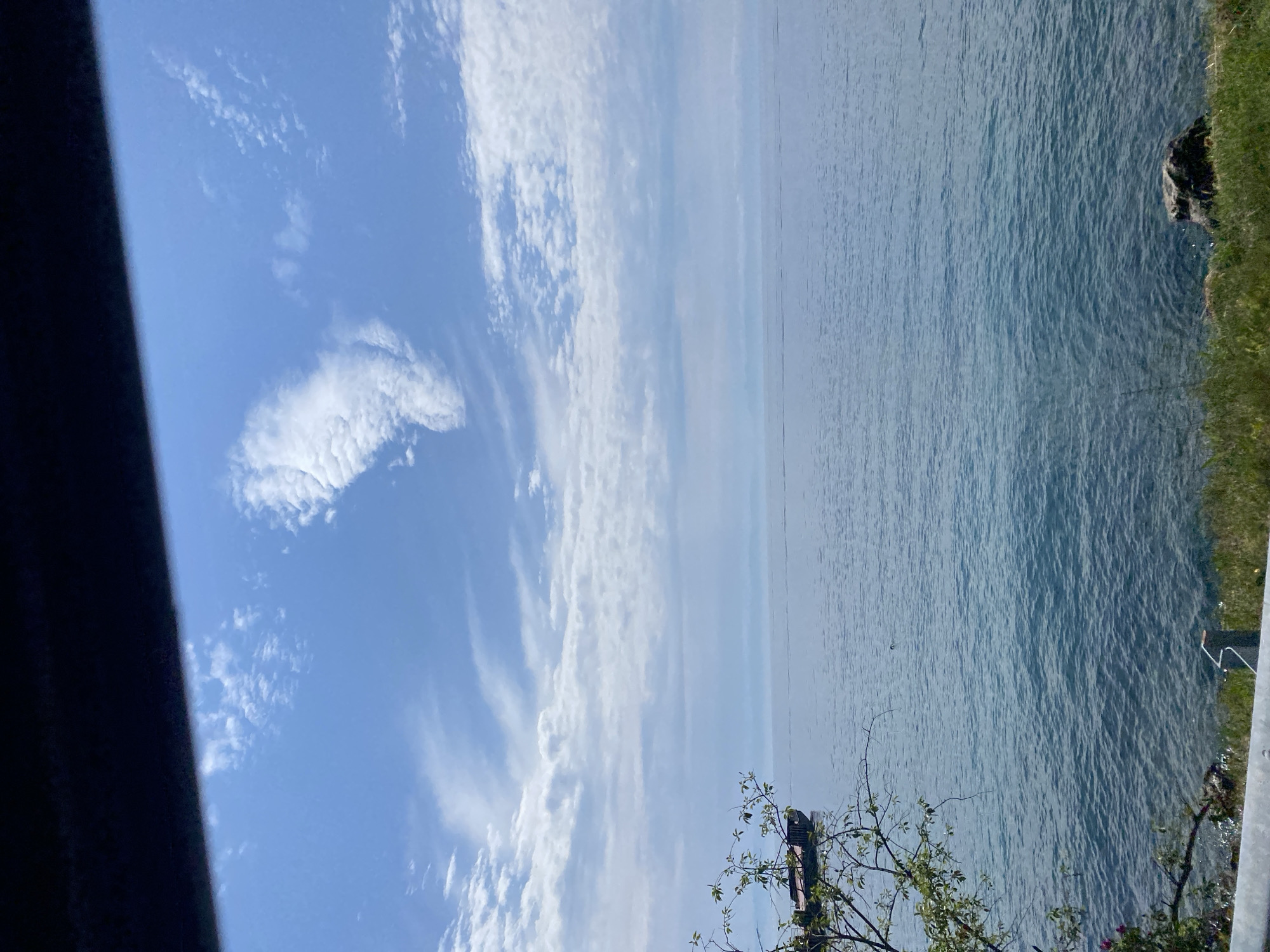 A serene scene unfolds overlooking a vast body of water, likely Lake Baikal, given the context of Listvyanka, Russia. The sky is a brilliant blue, dotted with wispy white clouds that stretch across the horizon. In the distance, faint landforms are visible, shrouded in a soft haze. The water is a deep blue-green, with gentle ripples disturbing its surface.
To the left of the frame, a small portion of a wooden boat is partially visible, moored near the shore. The boat appears weathered, hinting at its age and use. In the foreground, a lush green grassy area meets the water's edge. A few rocks are scattered along the bank, and a glimpse of a metal railing suggests a viewing or access point. A single tree branch with green leaves extends into the frame from the left, adding a touch of natural beauty to the composition. The overall impression is one of tranquility and natural splendor.