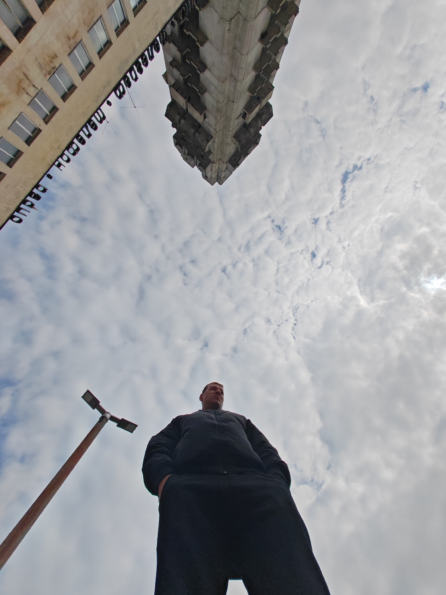 A low-angle photograph captures a male subject in the foreground against an overcast sky, framed by two prominent structures and a streetlight. The location is Shumen, Bulgaria.
The upper two-thirds of the image show a sky dominated by scattered cumulus clouds, with a brighter area in the upper-right corner indicating the obscured sun.
In the lower-right quadrant, a single male subject is visible from approximately the mid-torso down. He wears a dark, possibly black, zippered jacket and dark trousers, with both hands inserted into his pockets. His head is slightly tilted upwards and to the right, exhibiting a neutral expression.
On the left side of the frame, a multi-story building facade, light beige in color, is visible. It features several rectangular windows with reflective panes. Prominent Cyrillic text, reading "Дворец на културата 'Н. Вапцаров'" (Palace of Culture 'N. Vaptsarov'), is affixed vertically along its edge.
On the upper-right, a distinctive portion of the Monument to 1300 Years of Bulgaria (Founders of the Bulgarian State Monument) is visible. This structure consists of large, angular, grey concrete segments, characteristic of brutalist architecture, viewed from below.
In the lower-left quadrant, a reddish-brown streetlight pole stands vertically. It supports a horizontal crossbar with two modern, dark-colored light fixtures extending outward.
The scene depicts an outdoor environment with no other individuals or specific ground features visible beyond the subjects described.